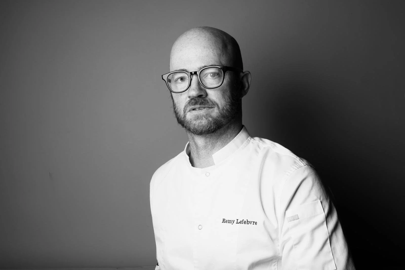 Black and white portrait of a man with glasses and beard in chef's uniform with the name "Remy Lefebvre" embroidered on it, standing against a plain background.