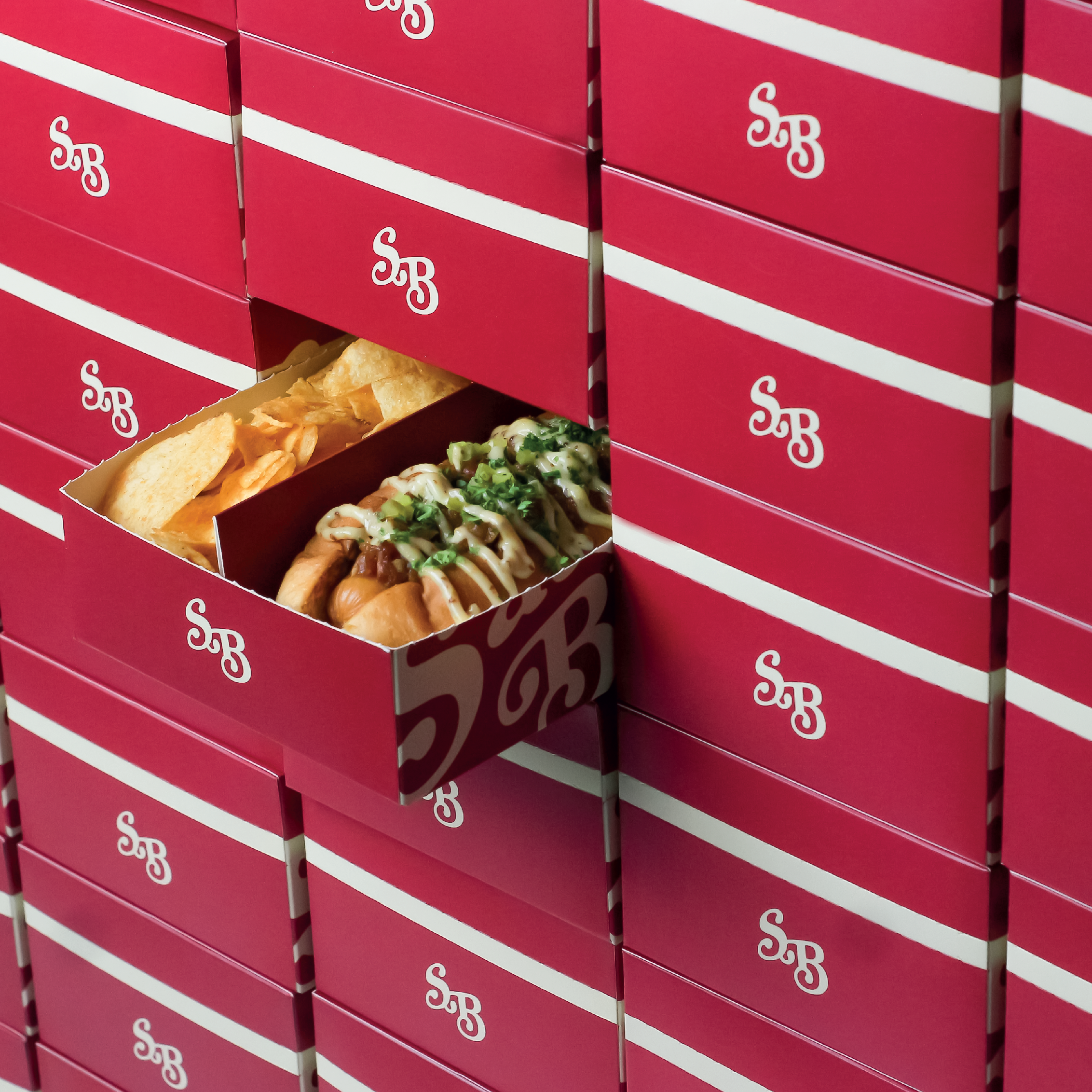 Open pink food boxes with white lettering, revealing hot dogs topped with green onions and sauce, and potato chips inside.