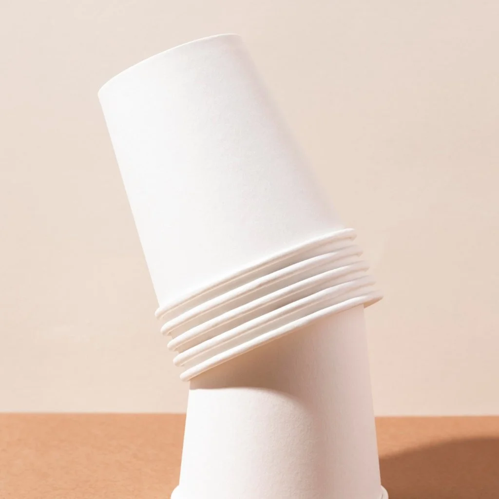 A stack of white disposable paper cups, with some cups tilted and overlapping on a wooden surface against a plain, neutral wall.