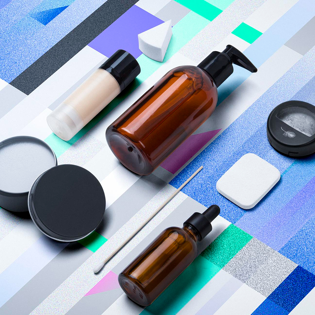 Beauty products, including two amber glass bottles with pump dispensers, a small powder puff, a black container with a closed lid, a cotton swab, a white sponge, and a small round black container, arranged on a colorful geometric background.