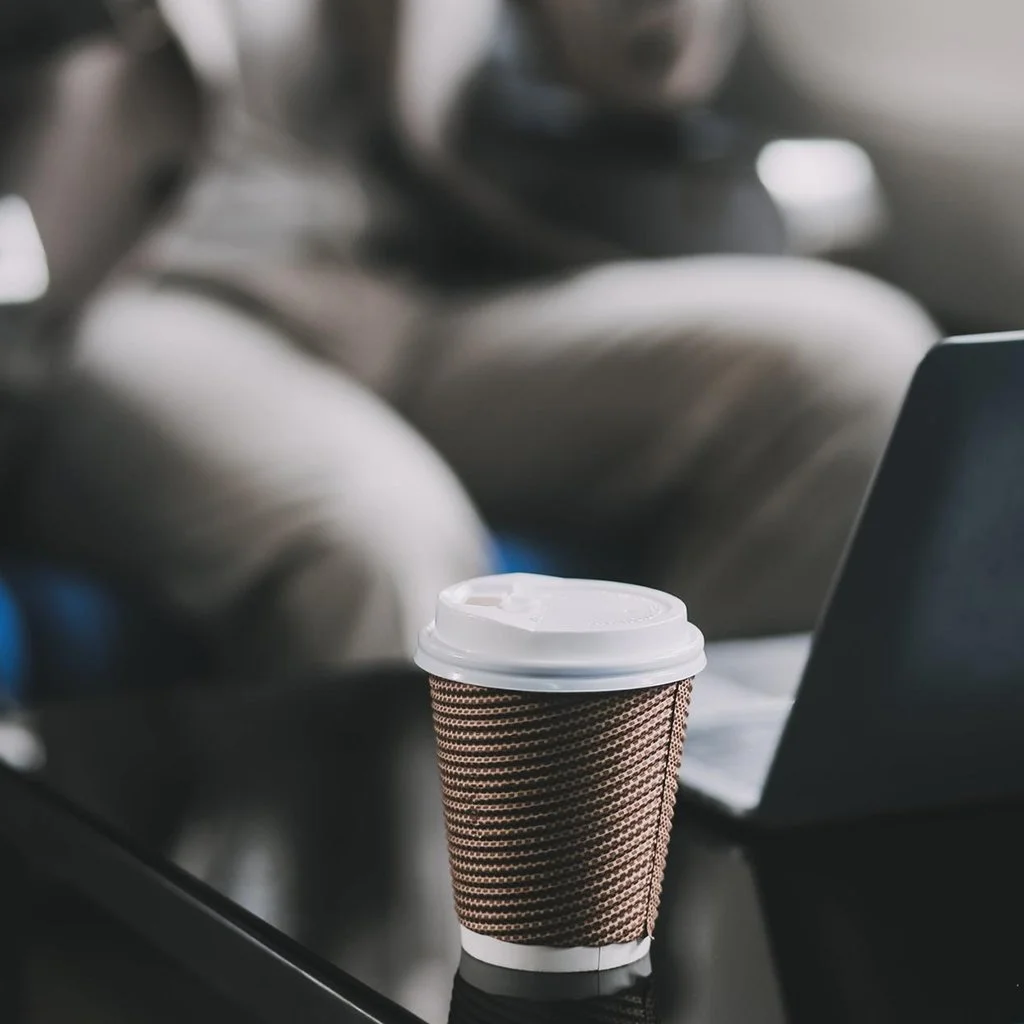 A disposable coffee cup with a brown patterned sleeve on a black table in front of a laptop in a cafe or airport setting.