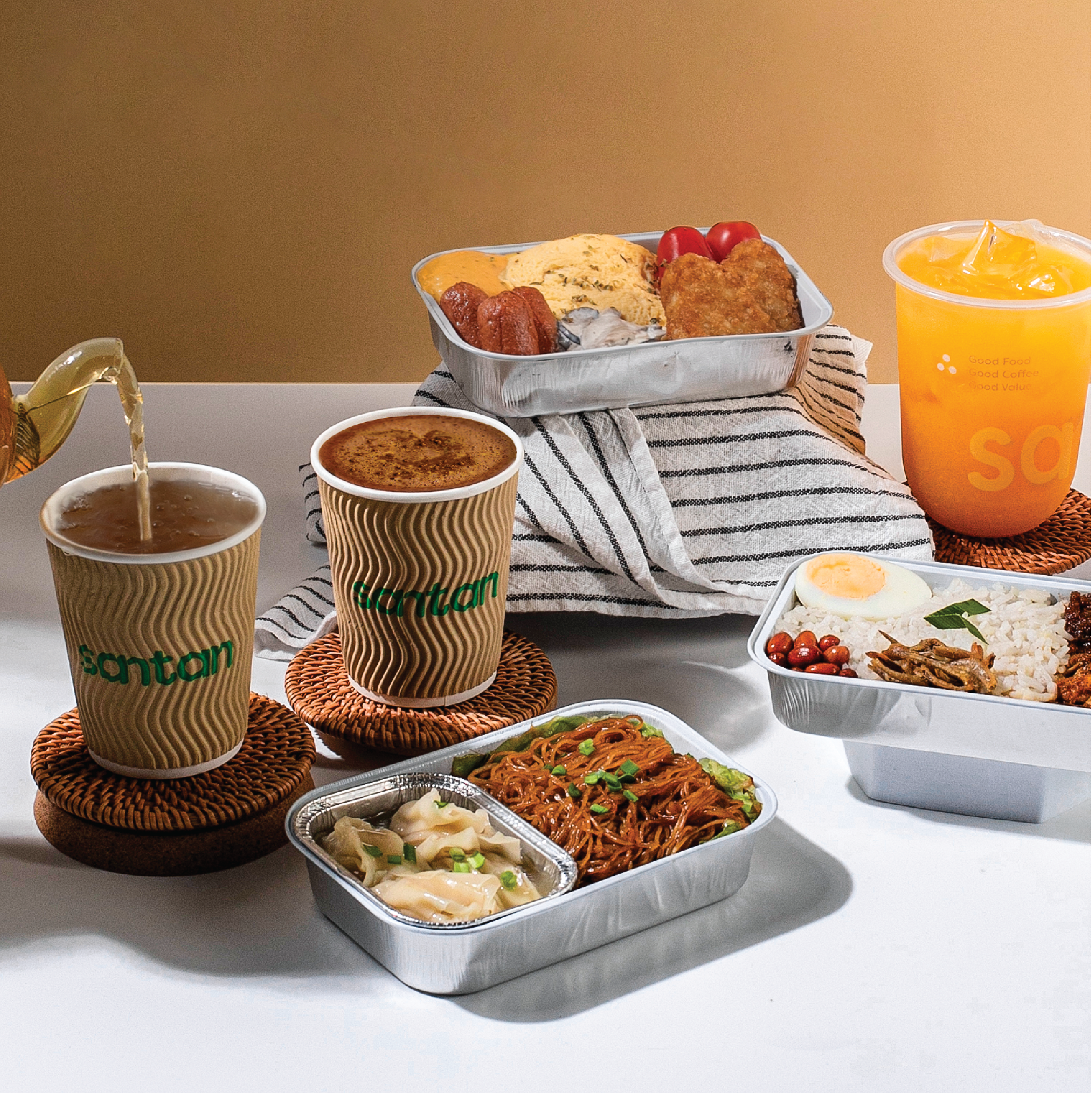 A spread of Asian-inspired takeout food and drinks on a white table, including fried rice with a boiled egg, stir-fried noodles, dumplings, a salad, two cups of tea, and a cup of juice, with a striped cloth and a metal tray of breakfast in the background.