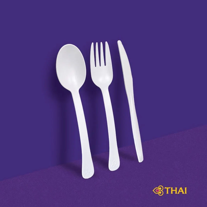 A white plastic spoon, fork, and knife arranged upright on a purple surface with a matching purple background, and the Thai airline logo in the bottom right corner.