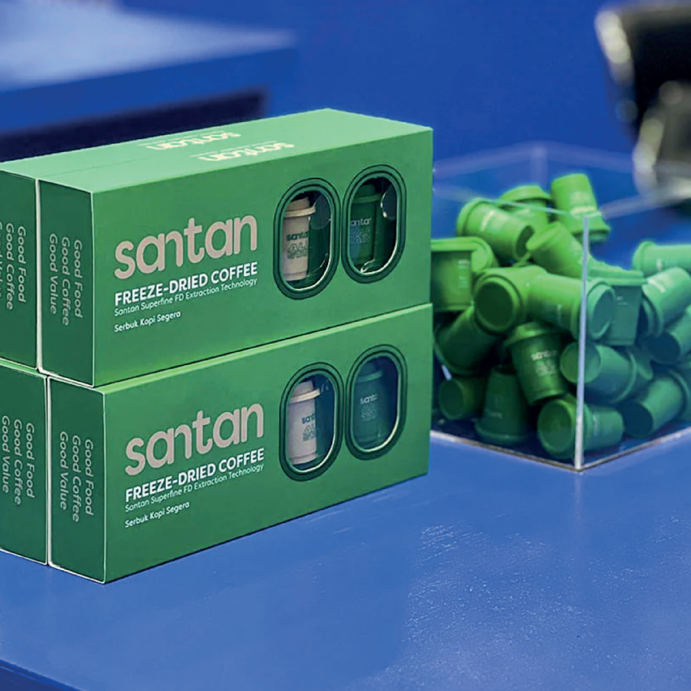 Green boxes of Santan freeze-dried coffee with small green capsules beside a container of more capsules on a blue surface.