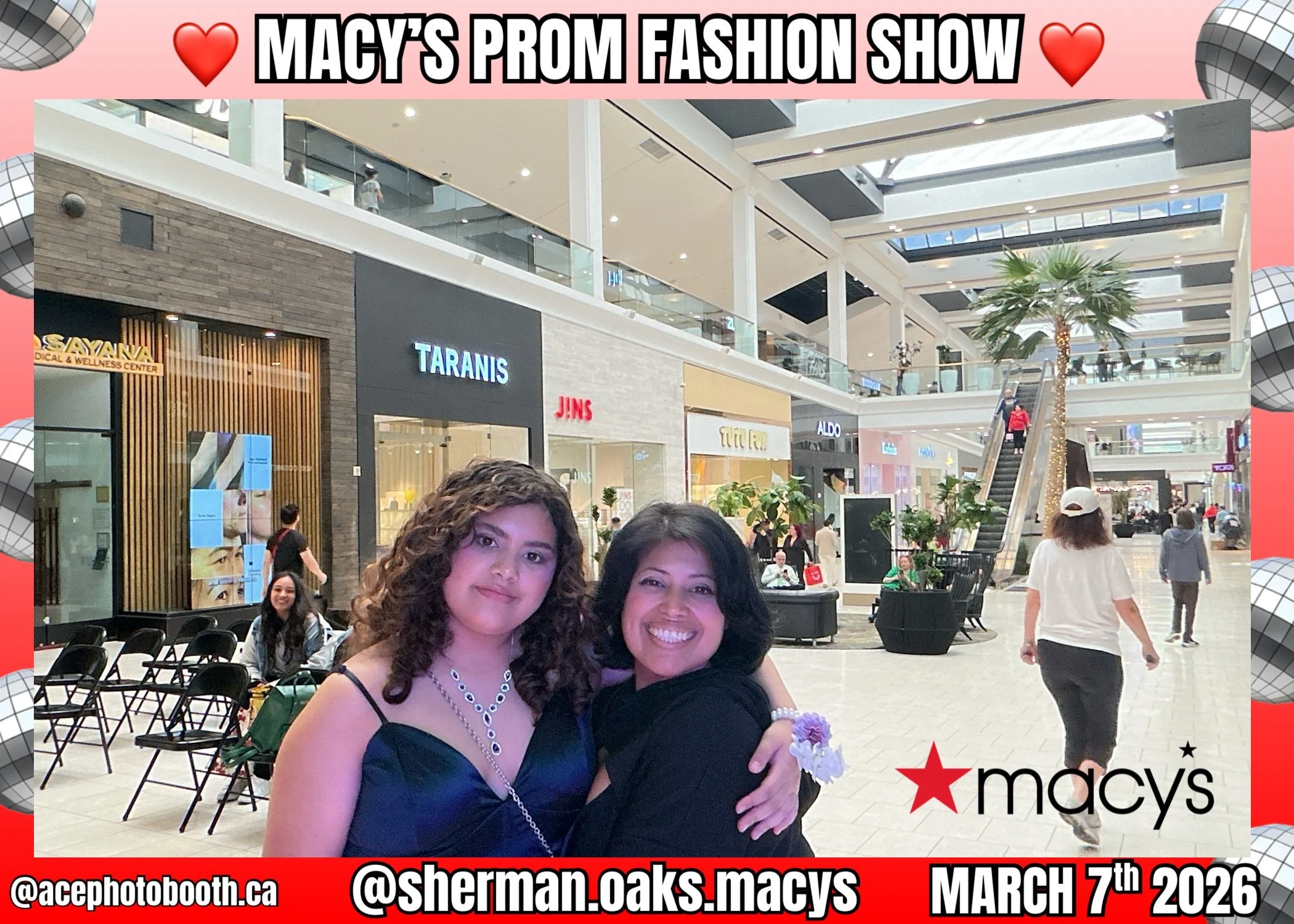 Two women smiling and hugging inside a shopping mall during Macy's prom fashion show. The mall has various stores, an escalator, and decorative plants, including a tall palm tree.