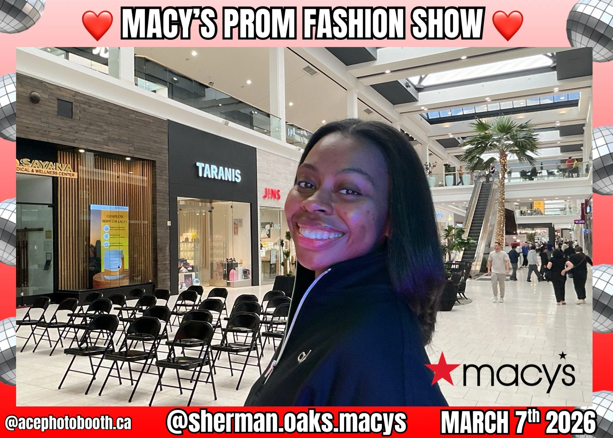 A smiling young woman with dark hair stands inside a shopping mall during Macy's prom fashion show in March 2026. She is in front of empty chairs and stores, with a large palm tree and multiple escalators in the background.