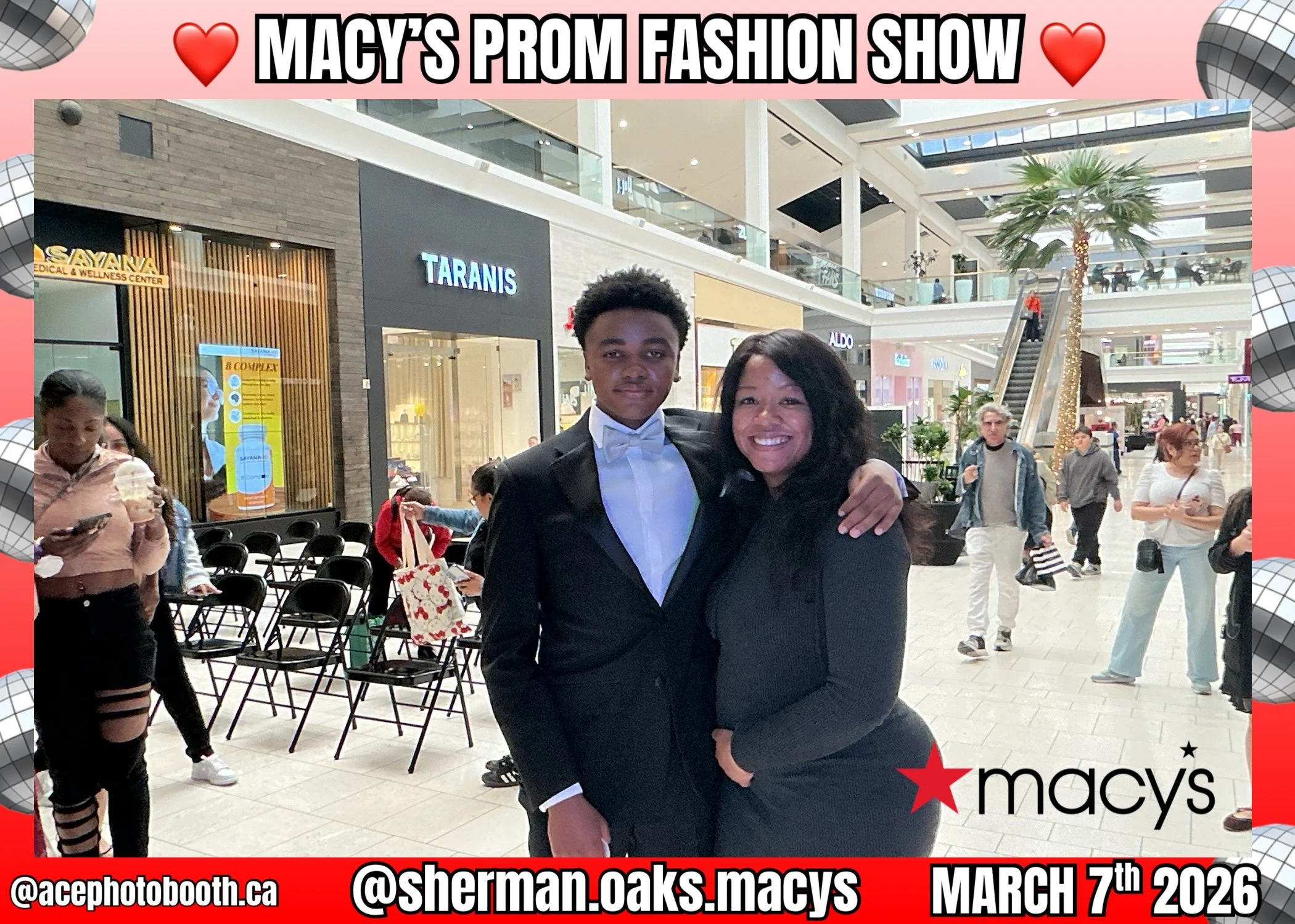 A young man in a tuxedo and a woman in a black outfit are posing together at Macy's mall for a prom photo. They are smiling and embracing, with mall storefronts and other shoppers in the background.