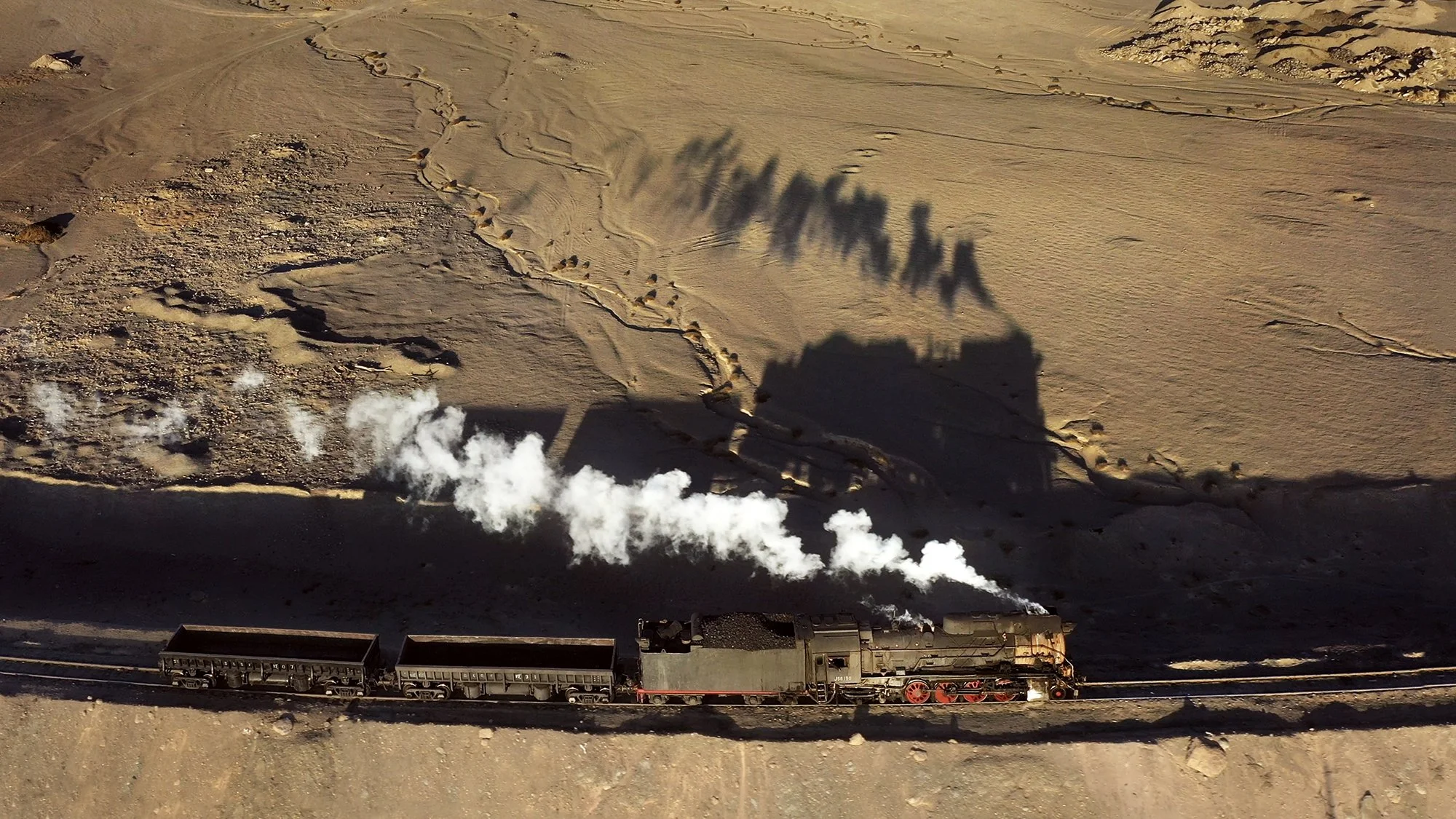  無人機視角下正在運行的蒸汽機車。機車和自翻車的影子映照在荒涼的土地上，2019年12月 