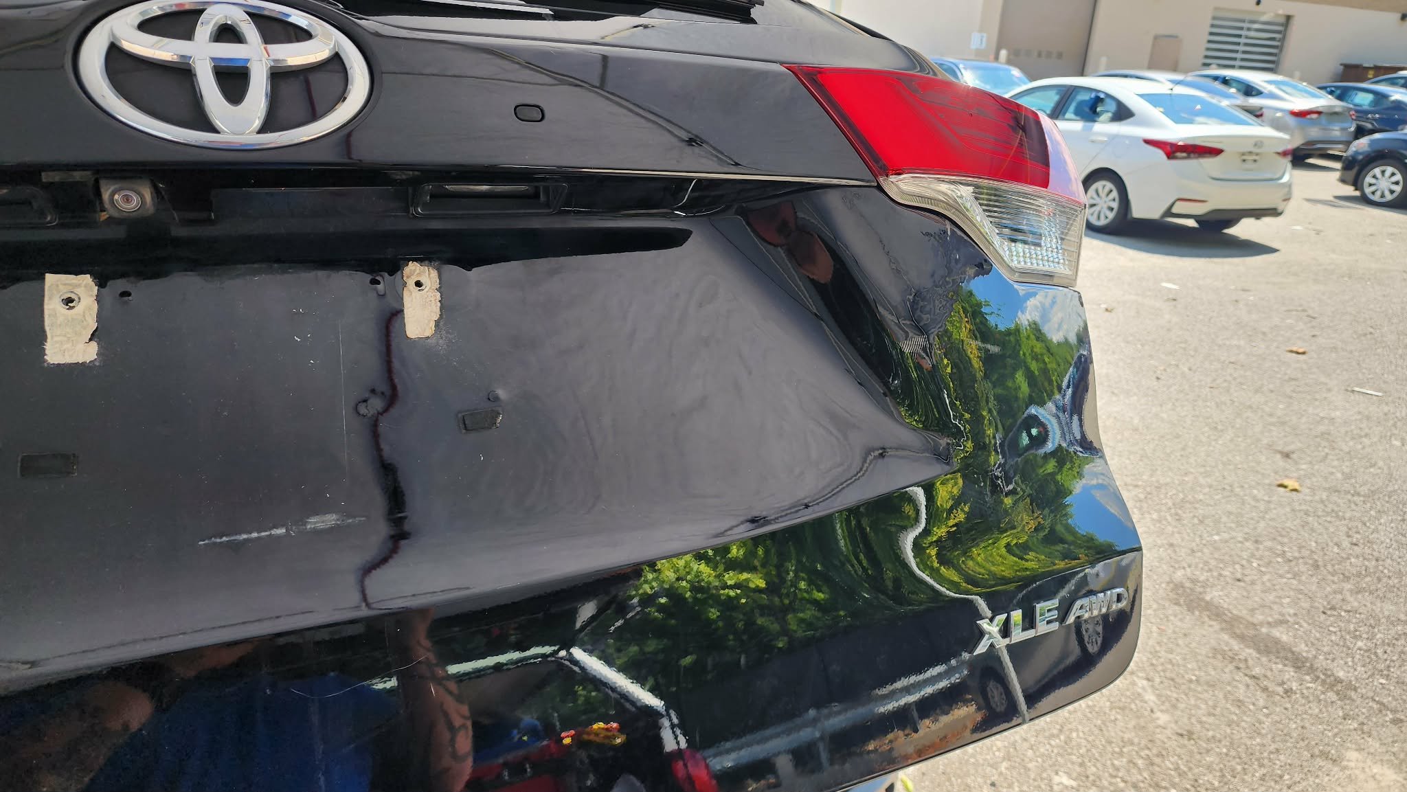 Rear of a black Toyota car with visible damage, missing license plate, and an XLE AWD badge in a parking lot with several other cars.