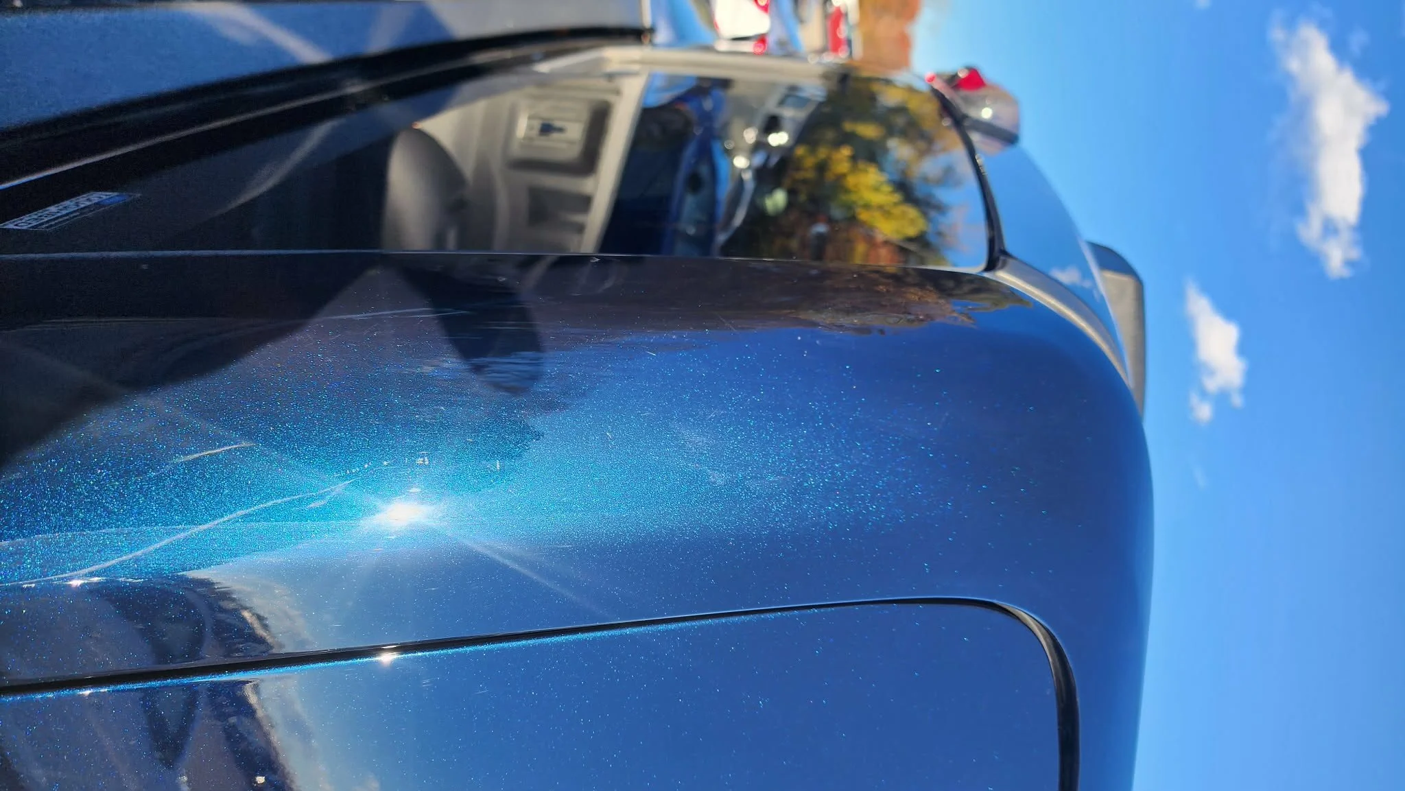 Close-up of a shiny blue car's front hood with reflections of nearby vehicles and trees, under a partly cloudy blue sky.