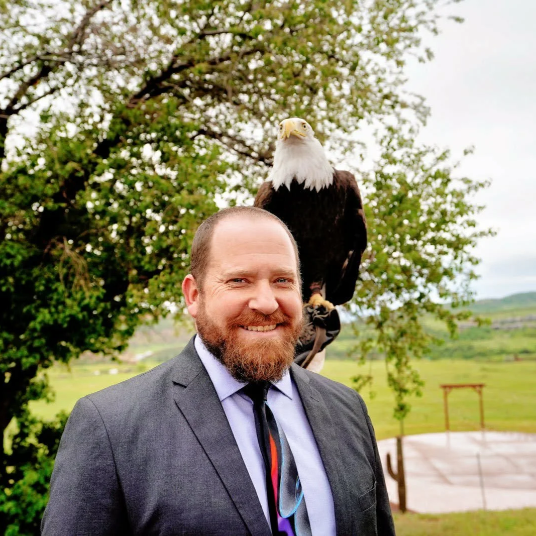 A man with a beard and short hair, dressed in a suit, smiling outdoors with a bald eagle perched on his shoulder, against a backdrop of green trees and grass.