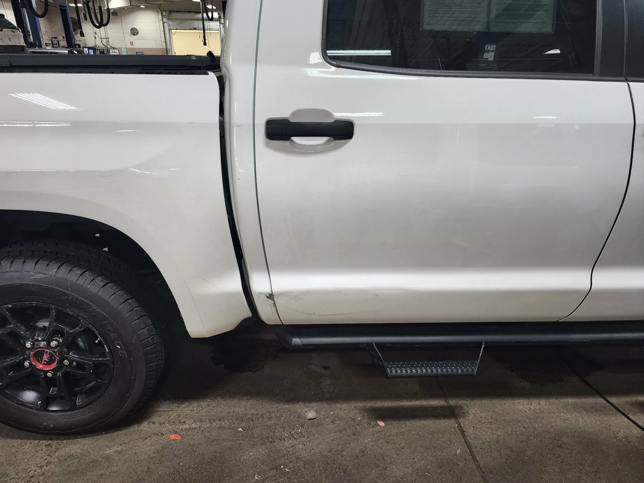 White pickup truck with black wheels parked indoors, showing the driver's side with visible damage and scratches on the door and fender.