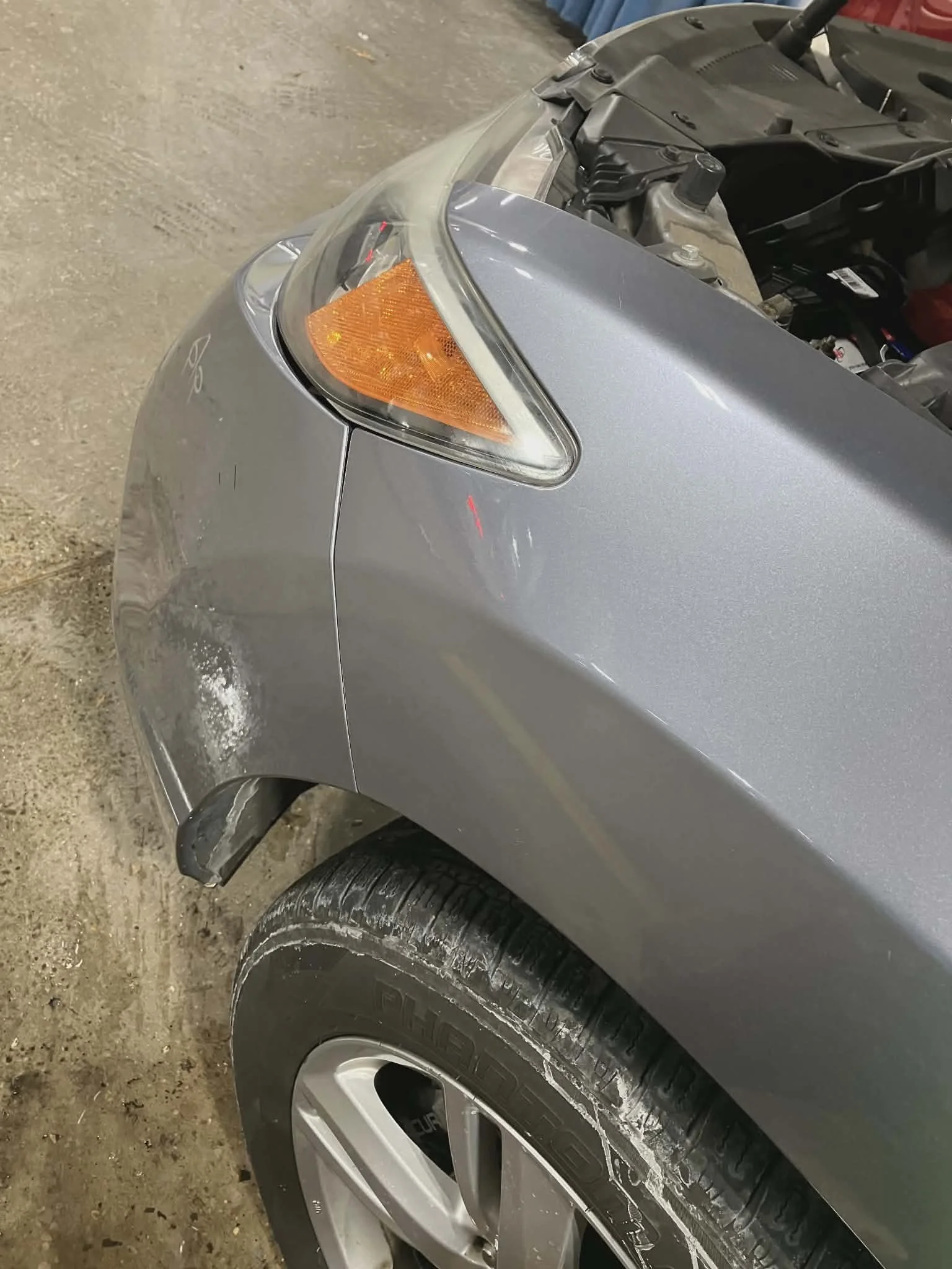 Front left side of a gray car with visible damage and dirt on the bumper and tire, in an indoor garage.