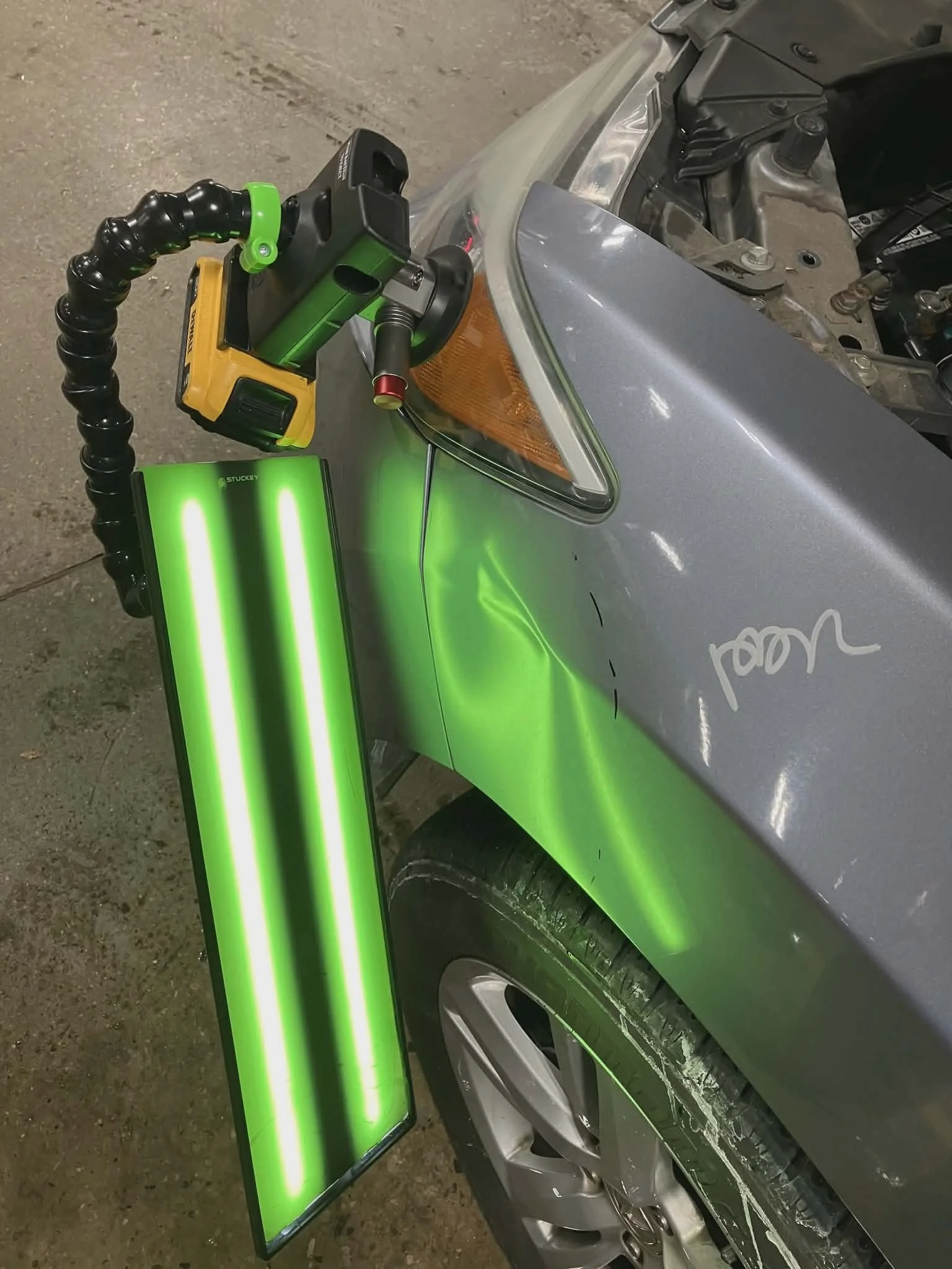 A silver car in a garage being repaired with a green and black robot arm holding a yellow cordless tool, a green portable light, and a bright green fluorescent light panel.