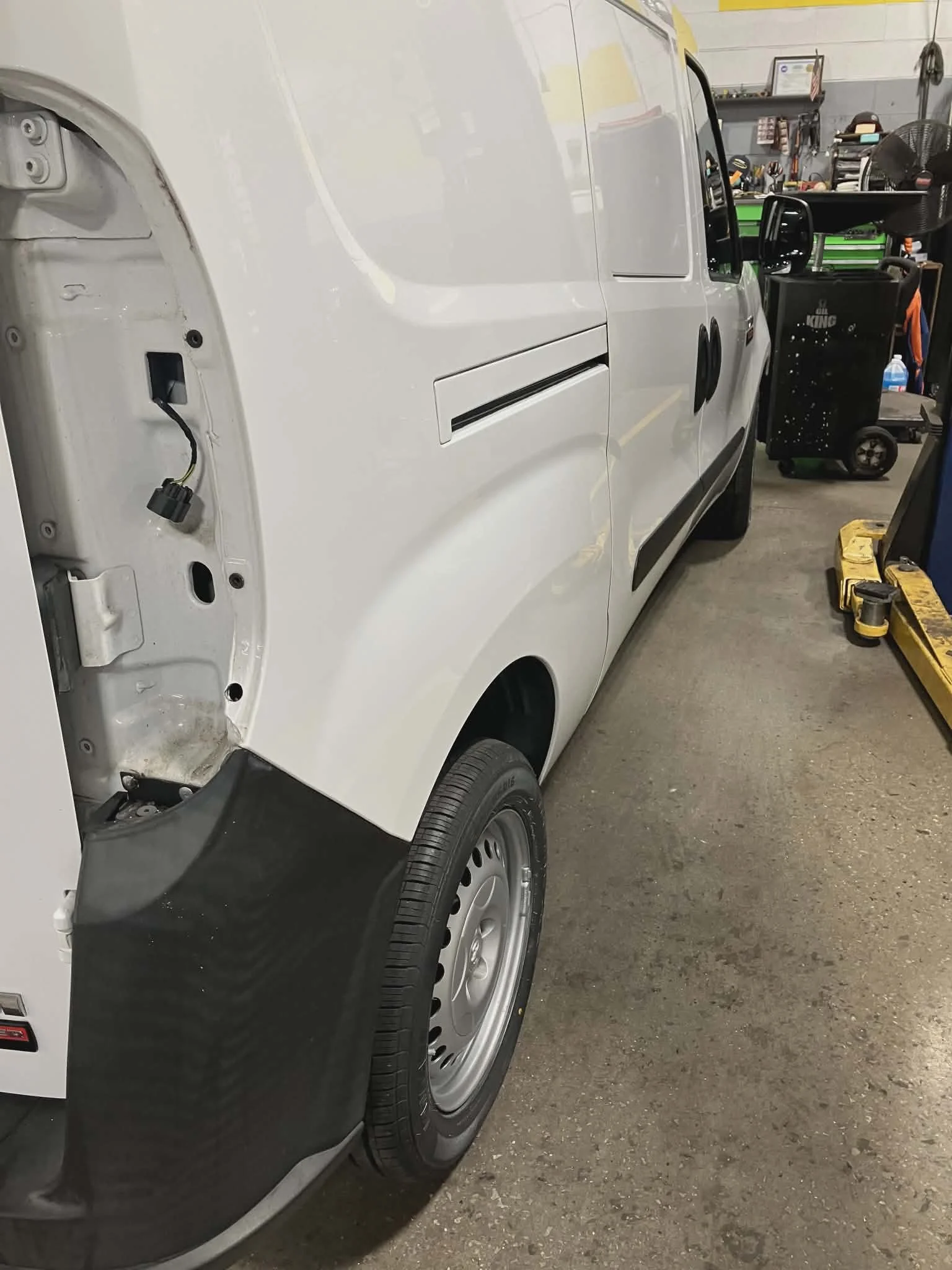 A white cargo van is in a garage, with the front left side missing its headlight and black bumper cover, revealing internal parts. The van is partially assembled, with a visible wheel and an area on the side door that appears to be for a handle or lo