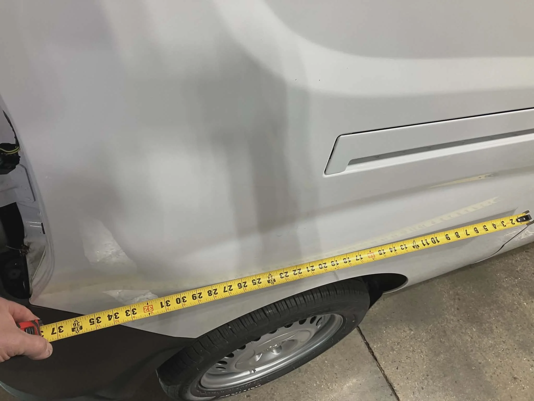 Front side of a silver car with a black tire, a vent, and a tape measure stretched across the lower part showing measurement.
