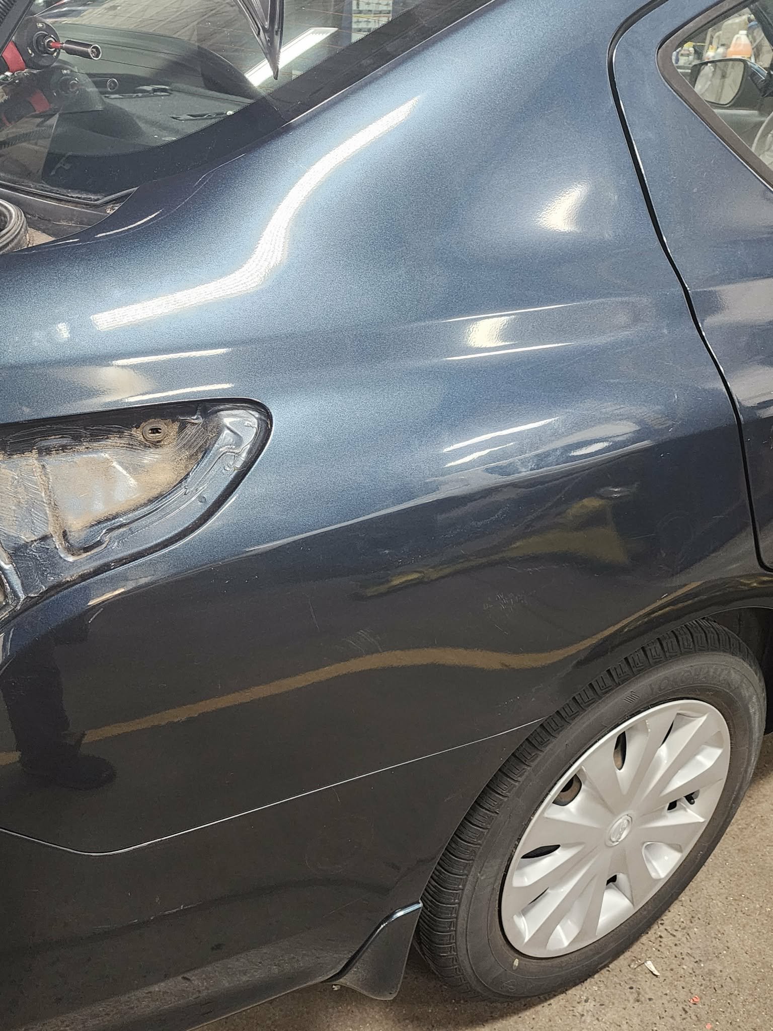The image shows the rear side of a blue car with a missing tail light cover, revealing internal components, and a clean black tire with a silver hubcap parked indoors.