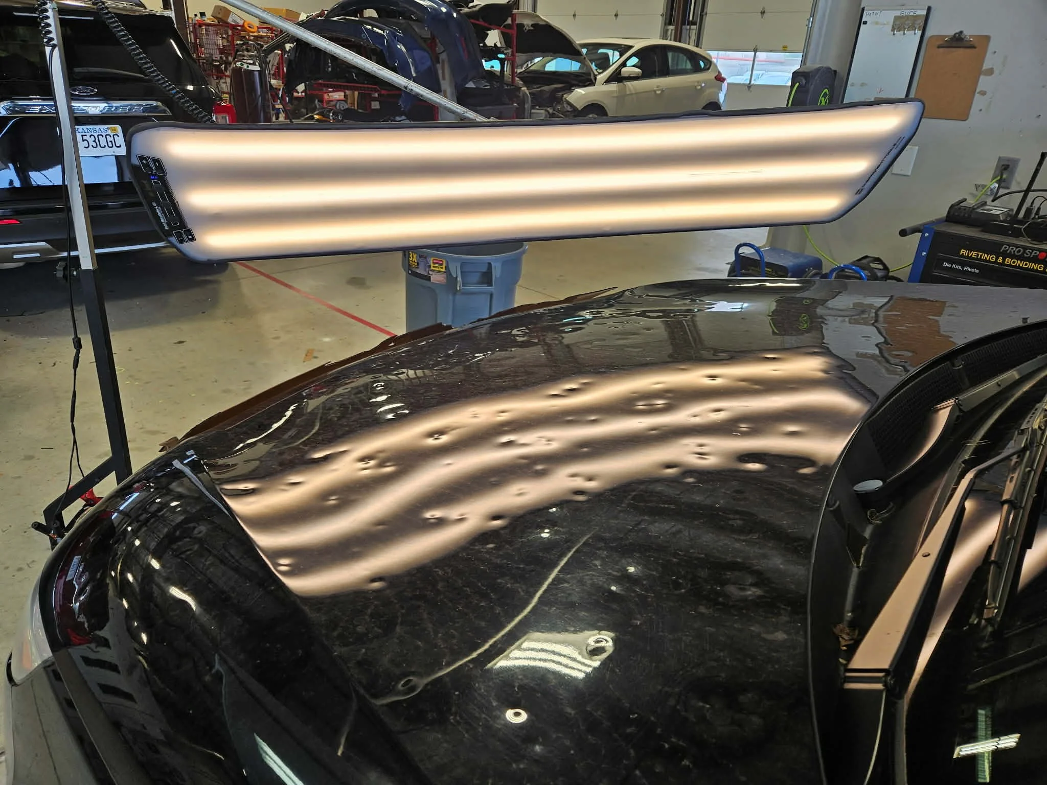 A black car with water spots and scratches on its hood, parked inside a garage. A large LED light is positioned above the car, illuminating the surface.