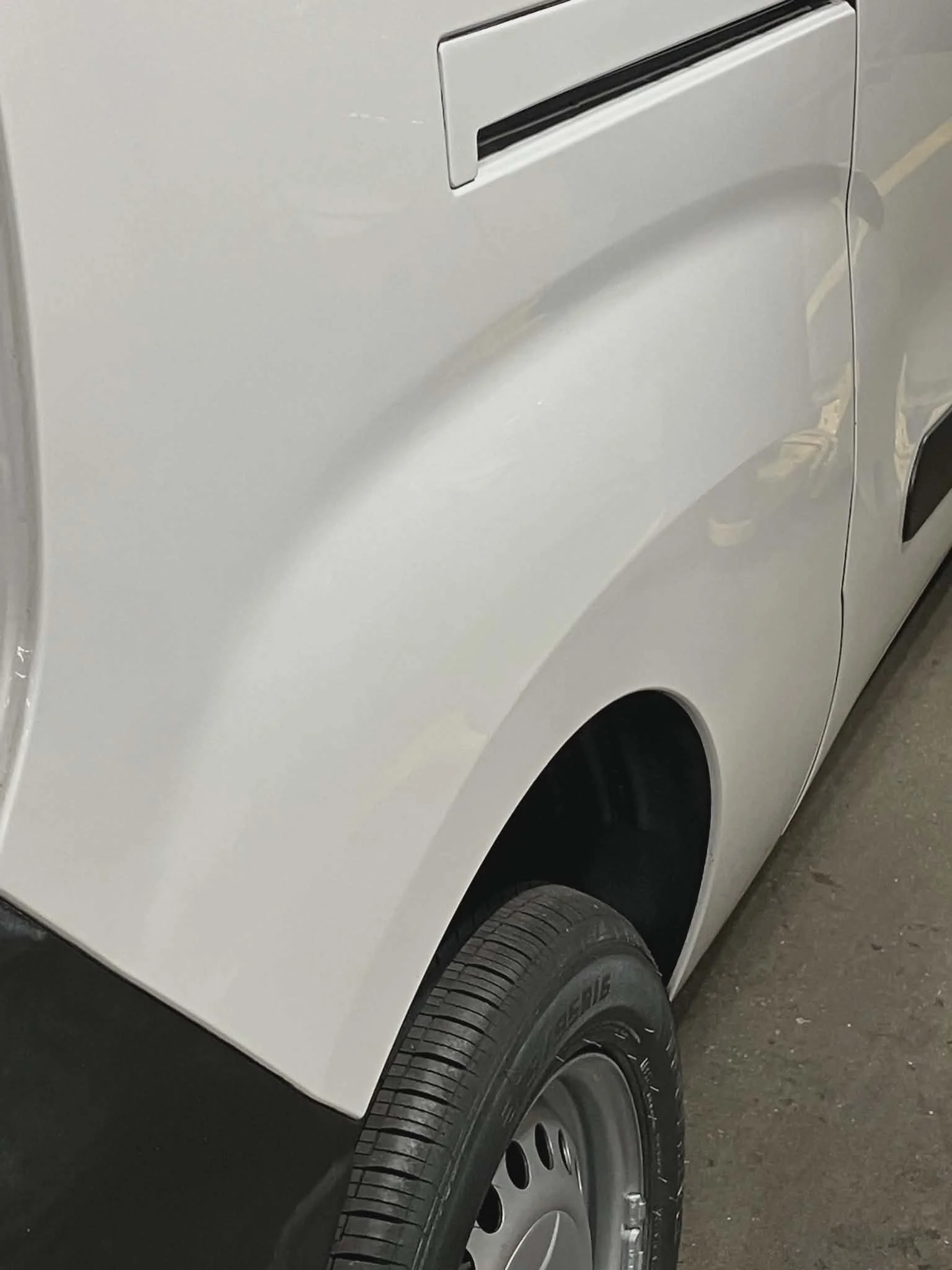 Close-up of a white vehicle's rear side, showing part of the rear panel, tire, and door handle.