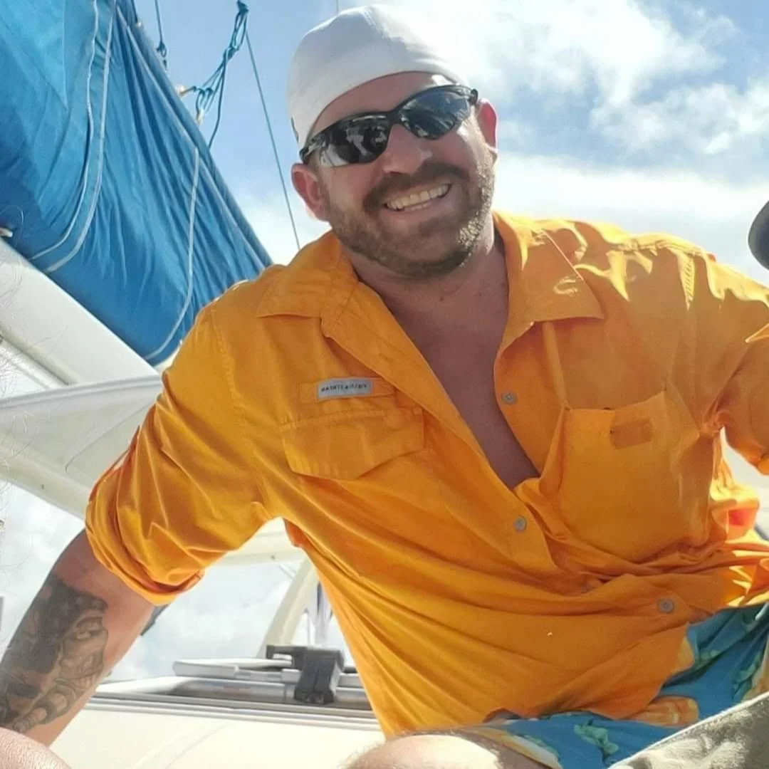 Smiling man wearing sunglasses, white cap, and yellow shirt on a boat with a blue sail in the background.