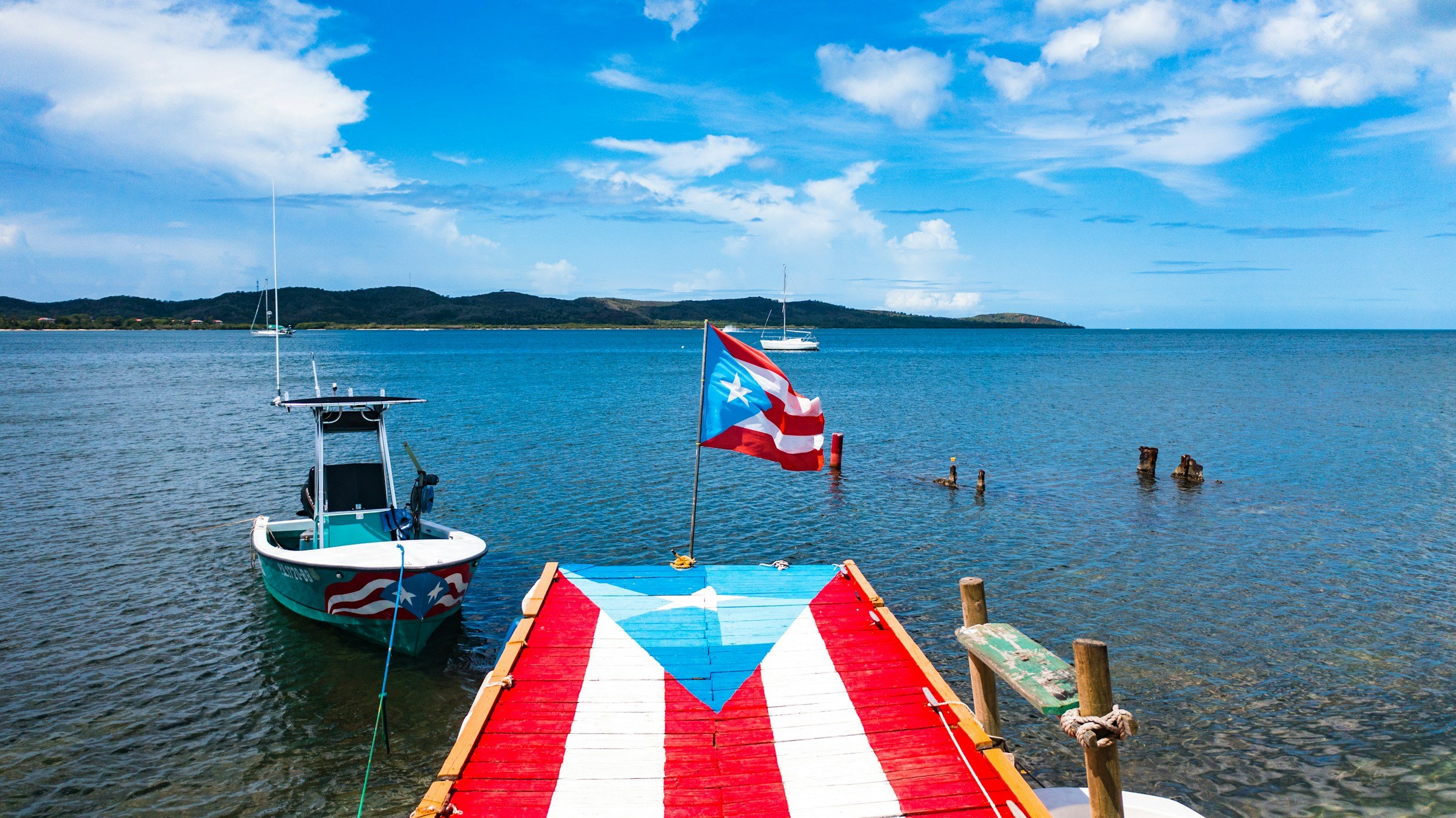 Puerto Rico flag on a small dock leading to boats in a calm bay with a mountainous island in the background and partly cloudy blue sky.