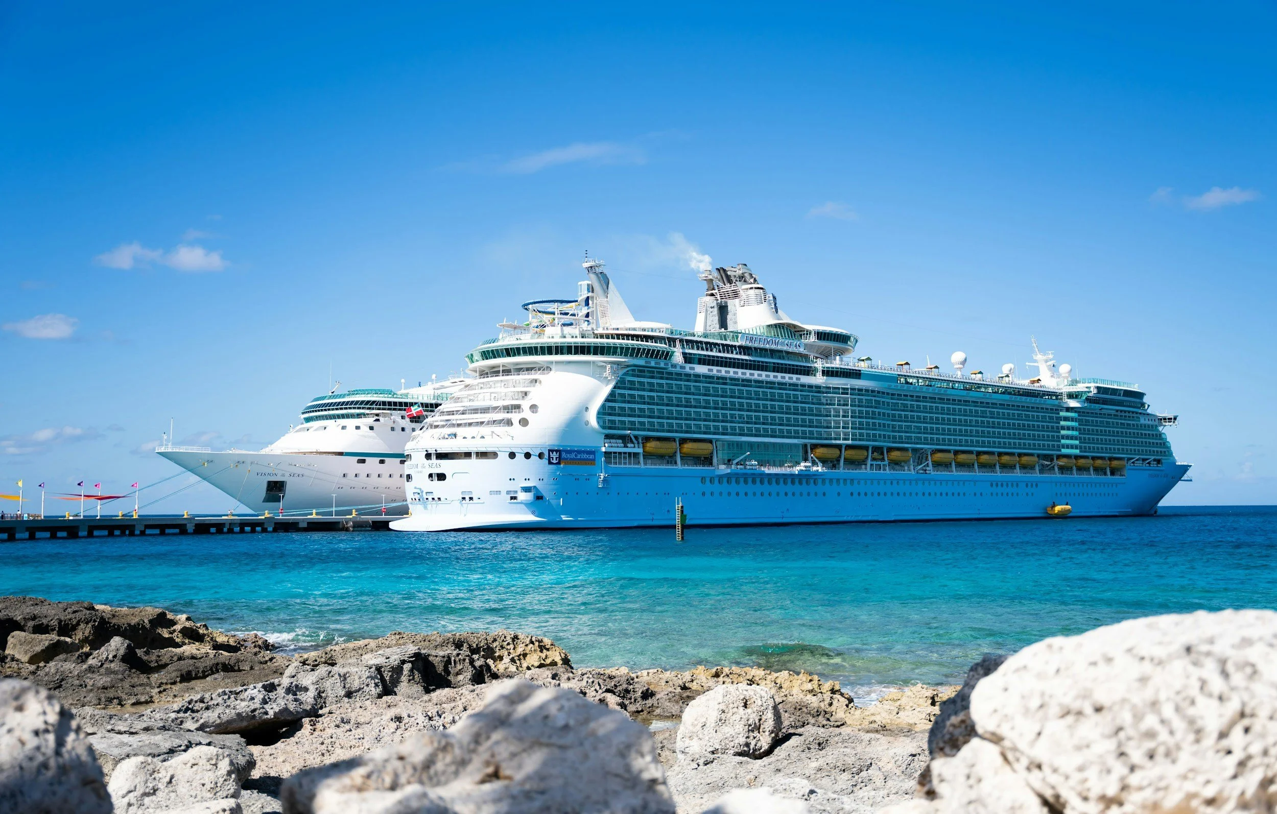 Two large cruise ships docked at a pier on a bright, sunny day with clear blue skies, rocky shoreline in foreground.