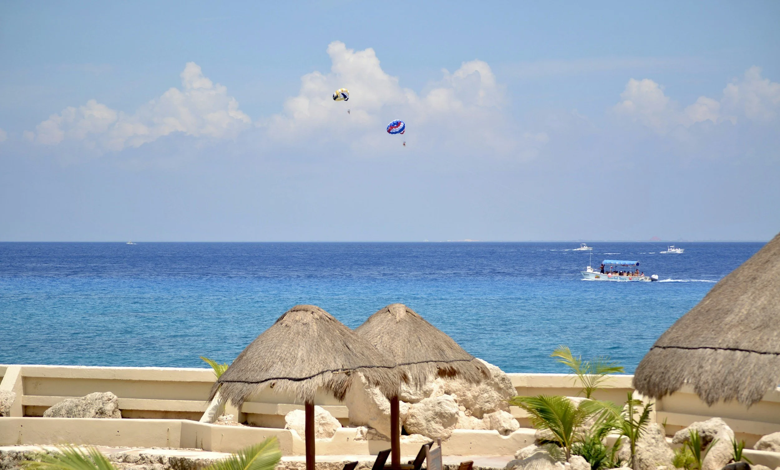 Beach scene with thatched umbrellas, blue ocean, and two parasails in the sky, with boats on the water.