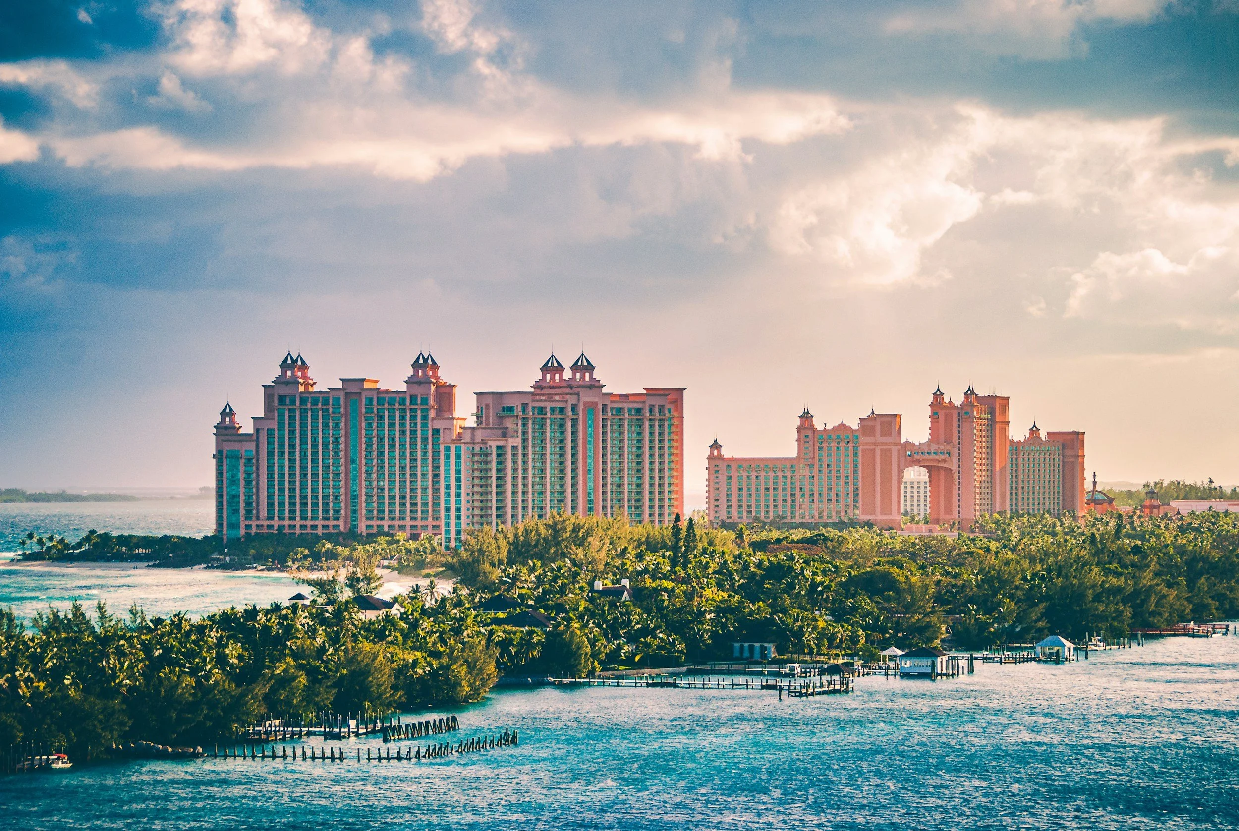A large hotel resort on the waterfront with multiple pink and blue towers and lush green trees in the foreground.