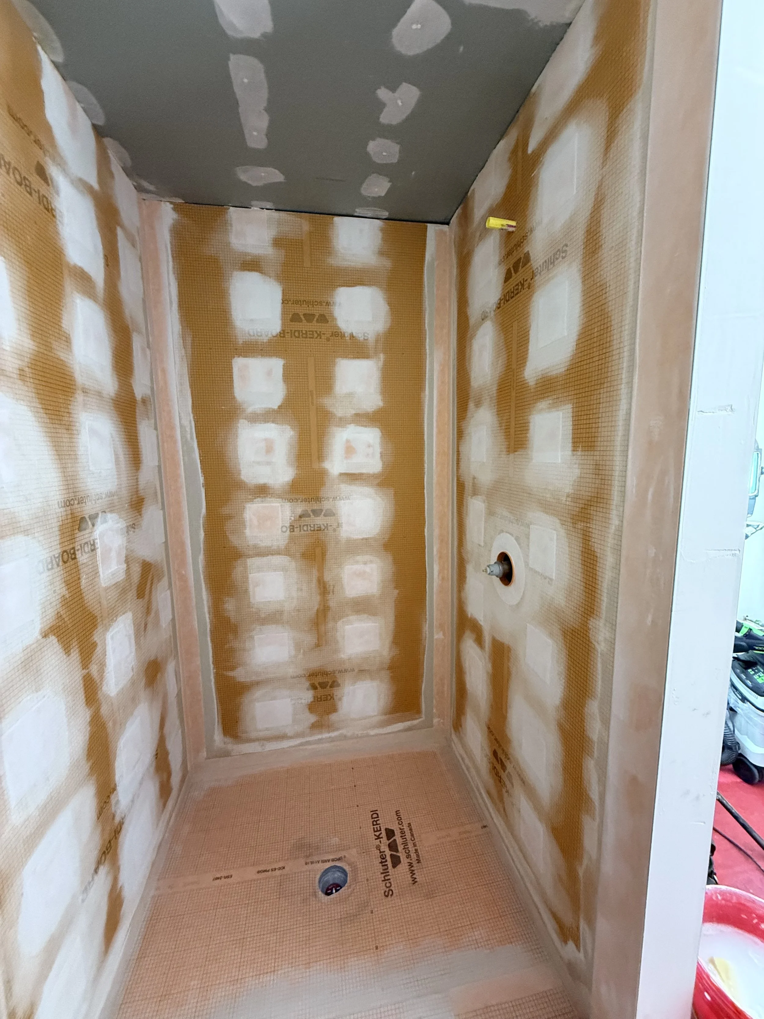 A small room under construction with partially finished drywall, a peg hole in the wall, and a floor drain. The walls and ceiling are patched, with some drywall tape visible.