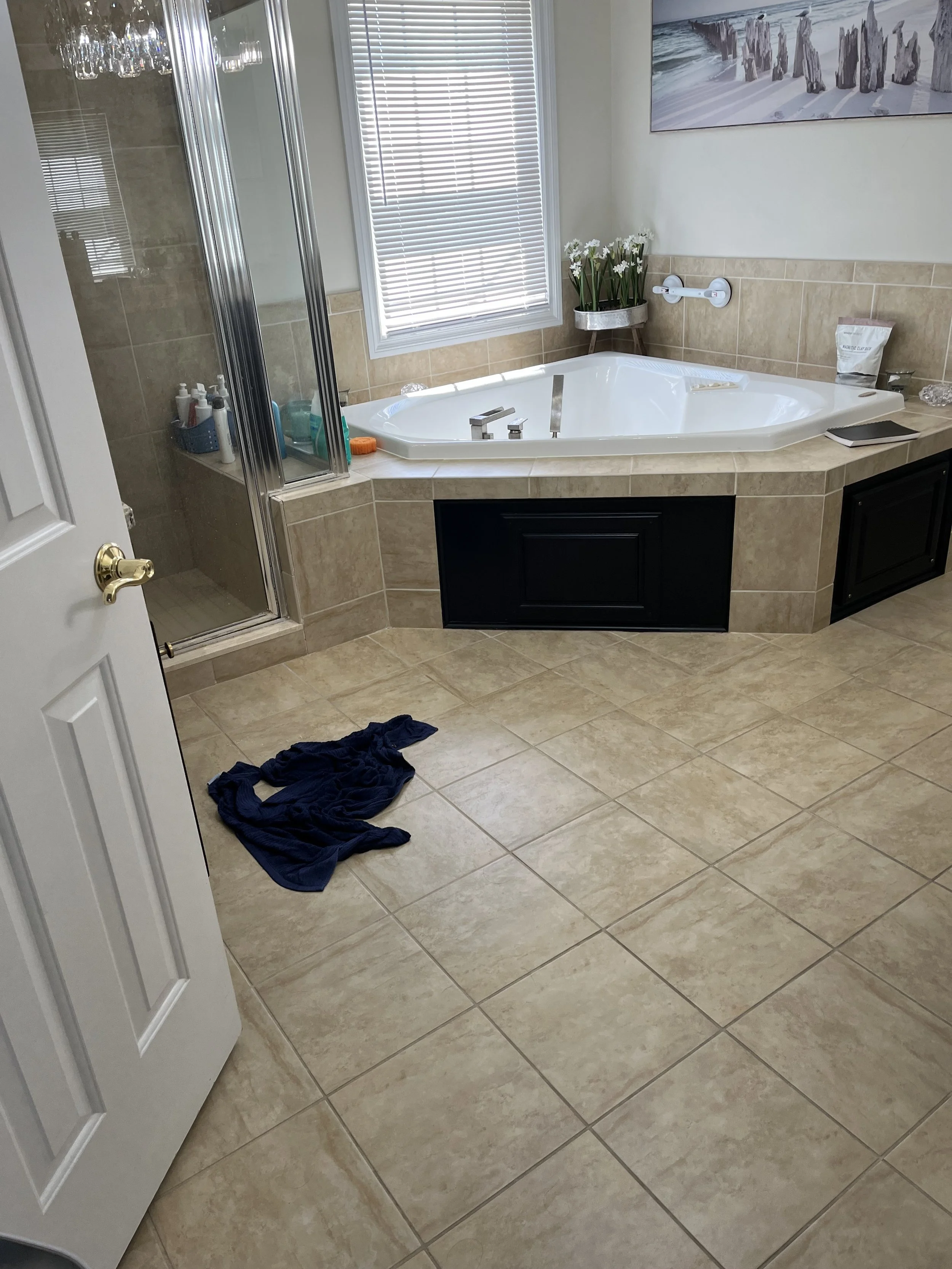 Bathroom with a jacuzzi tub, a glass shower, beige tiled floor and walls, a window with blinds, a small table with white flowers, a wall-mounted soap dispenser, and a towel on the floor.
