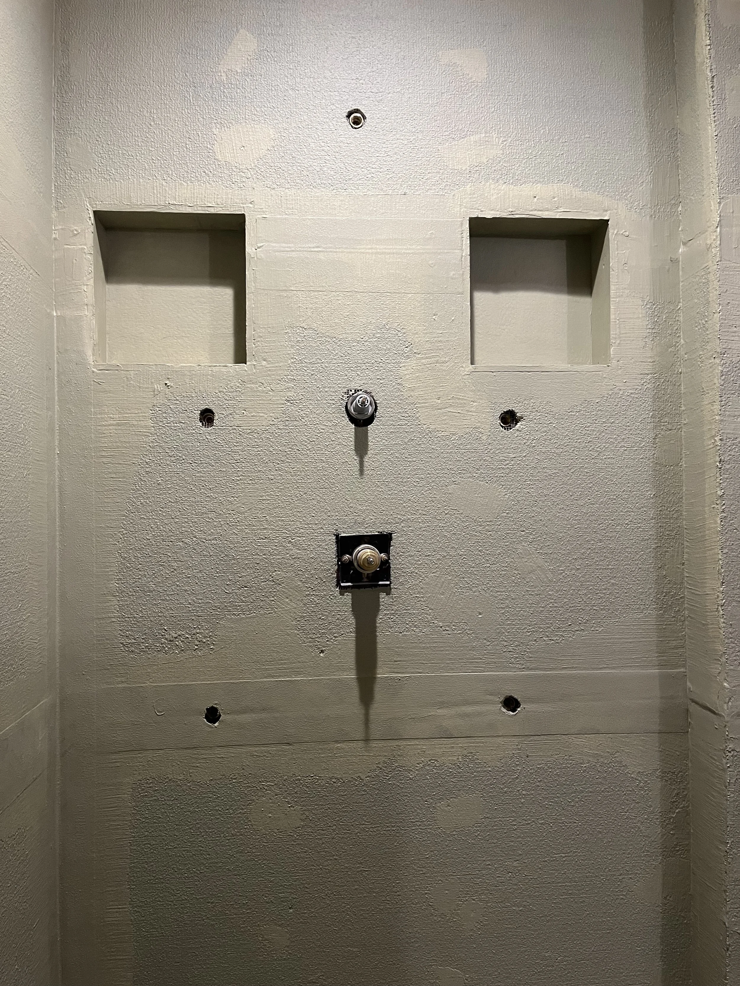 Empty wall with a single light fixture, several holes, two recessed wall niches, and a black plate with a round fixture hanging in the middle.