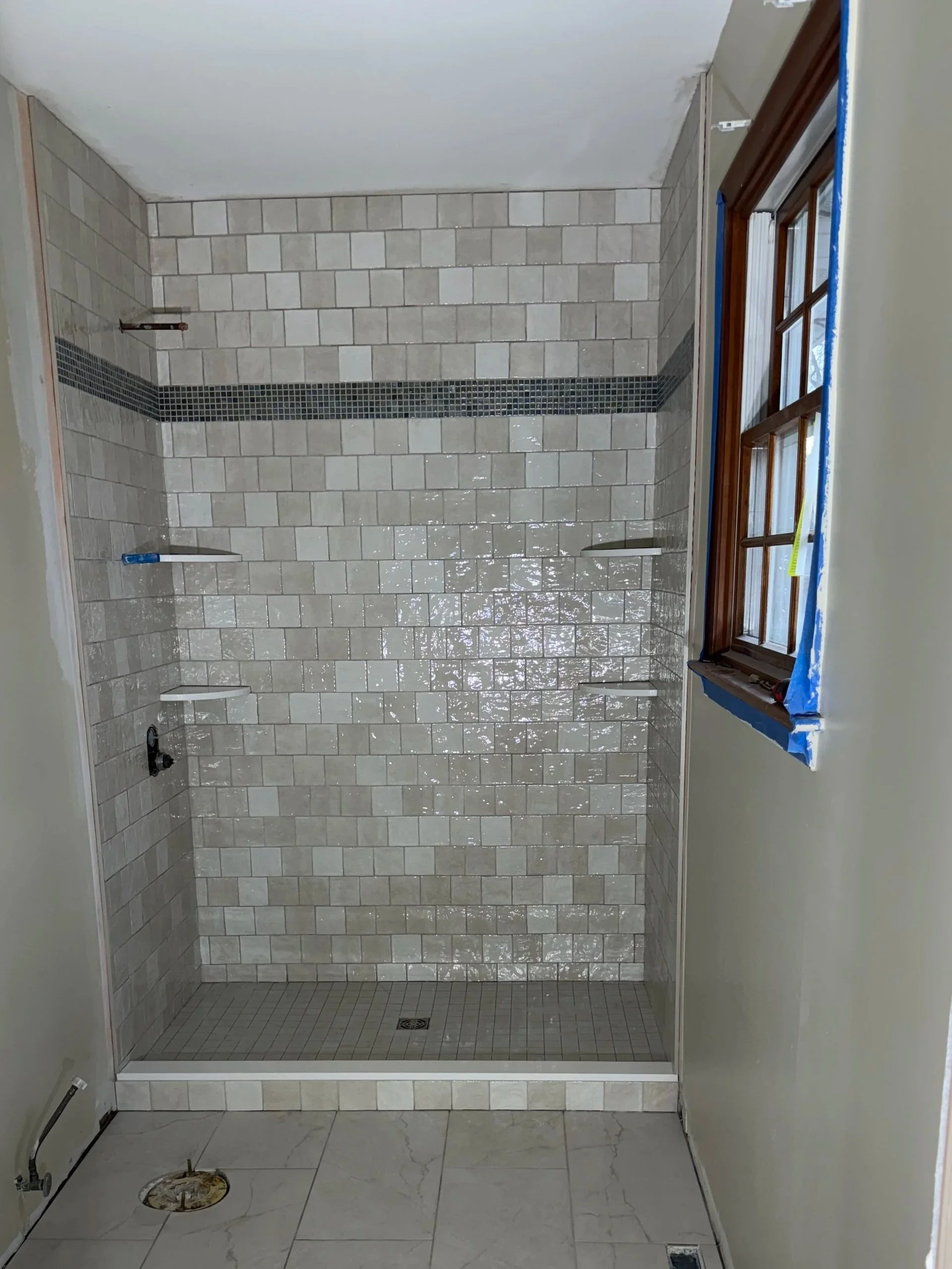 A newly tiled walk-in shower with beige and light gray tiles and a horizontal strip of small black tiles. The shower has multiple built-in shelves on both walls. The window next to the shower has a wooden frame and is partially covered with painter's tape. The bathroom floor is tile, and there is some plumbing work visible near the floor.