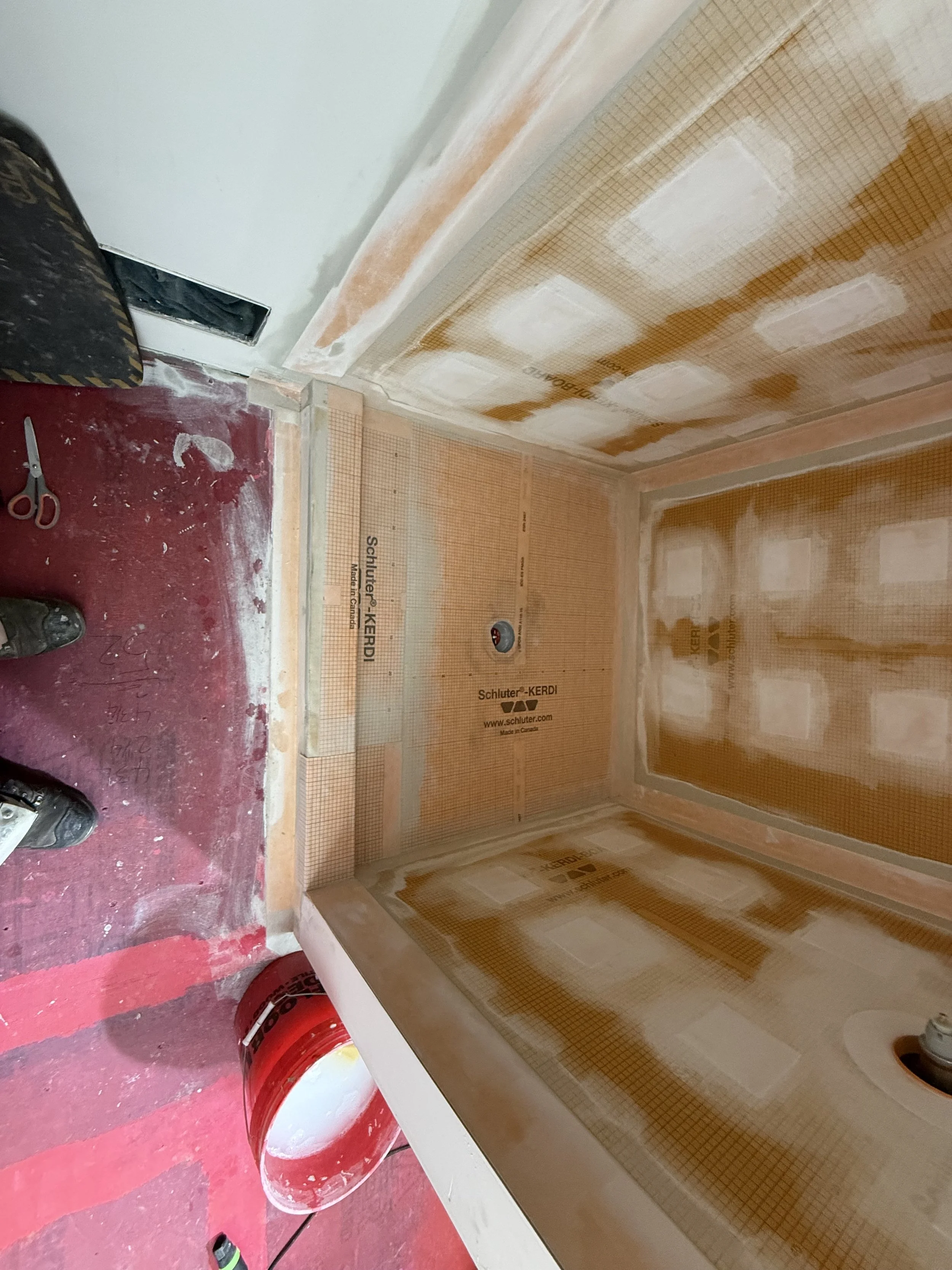 Interior of a bathroom under construction, with drywall installed, some areas taped and mudded, and a circular pipe opening in the floor for a toilet. Tools and construction materials are visible on the floor.