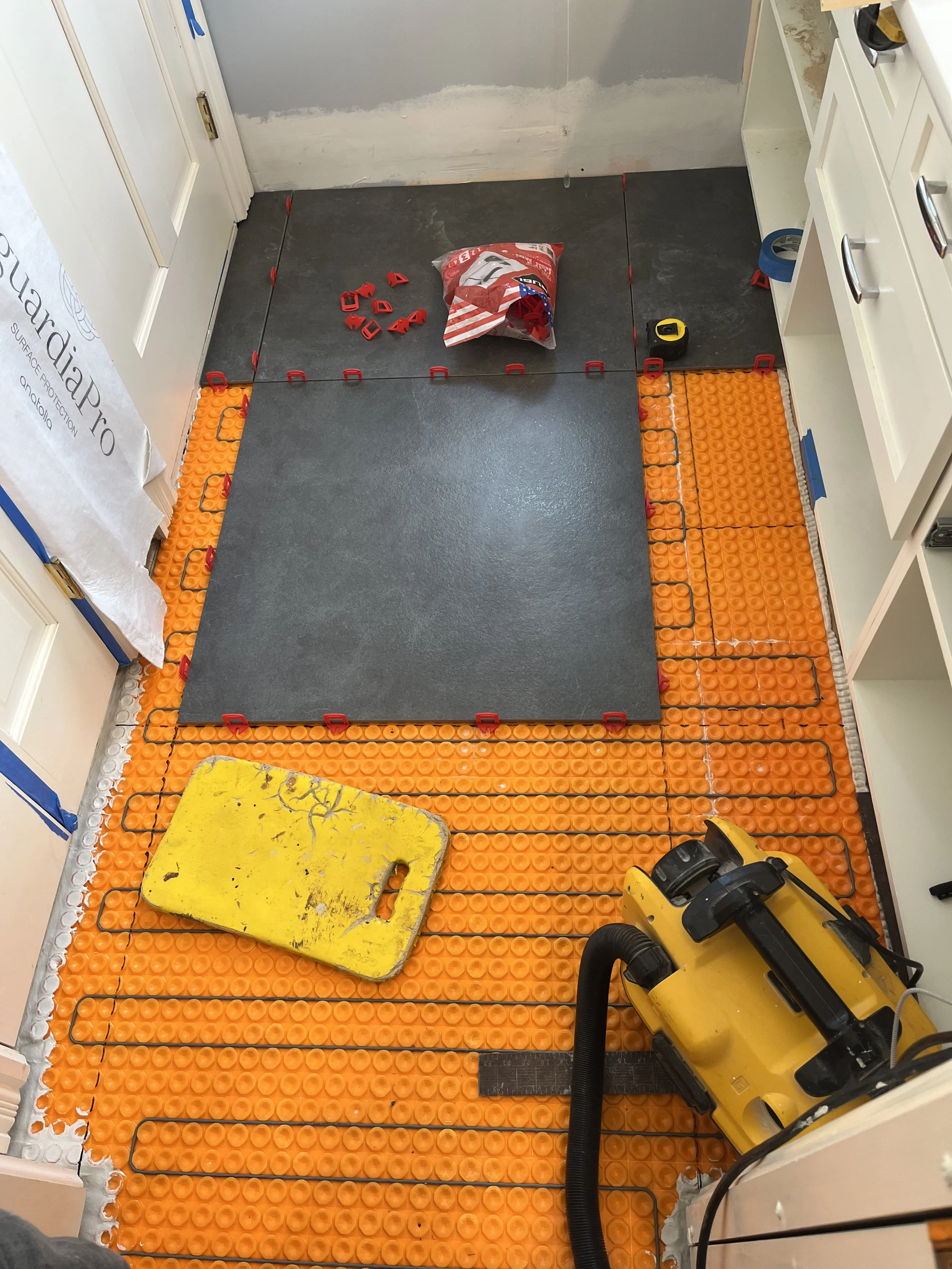 Bathroom floor being renovated with underfloor heating system installation. The floor is partially covered with orange heating mats, and a black tile is being installed in the center. Tools and materials, including a yellow push tool, a bag of tiles,