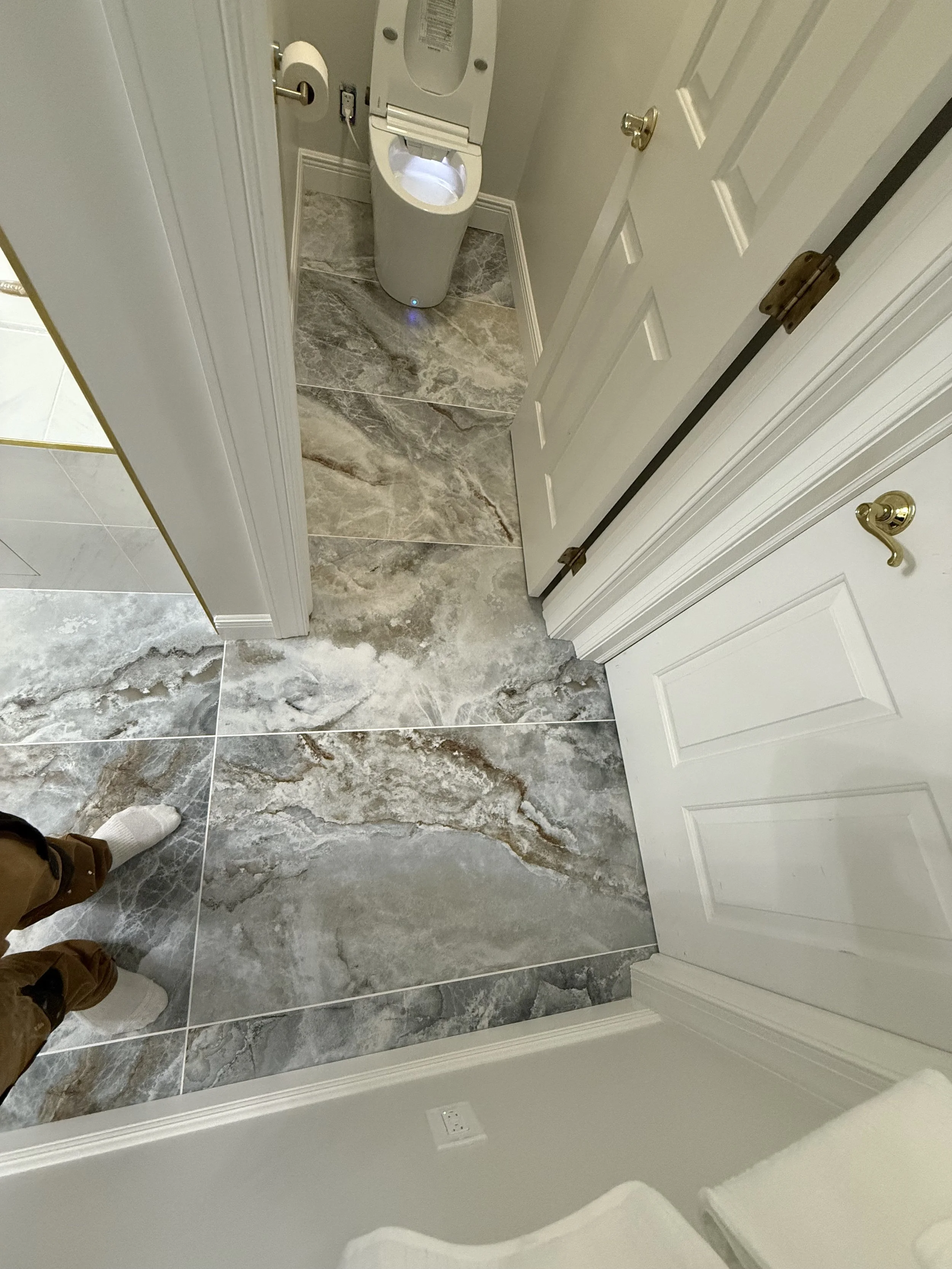 A bathroom with large marble tiles flooring, an open toilet, a closed white door, and a person's legs wearing brown pants and white socks visible in the mirror reflection.