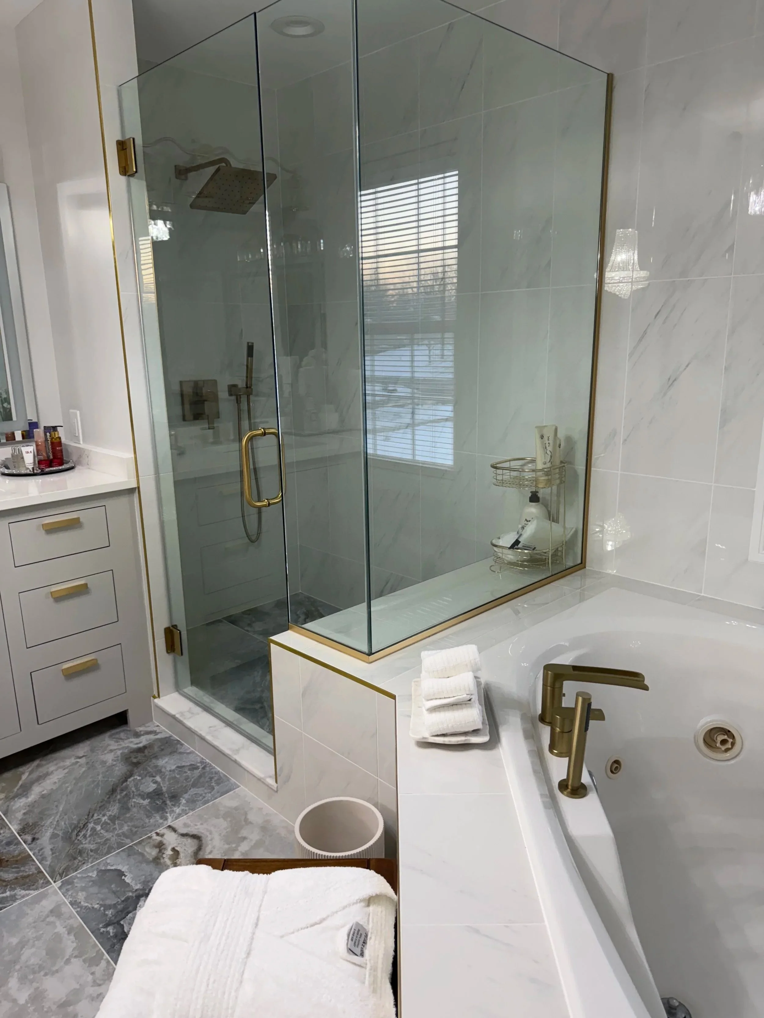A modern bathroom with a glass-enclosed shower featuring gold fixtures, a bathtub with a gold faucet, and a white cabinet with gold hardware. There are towels, a small trash can, and a window with blinds.