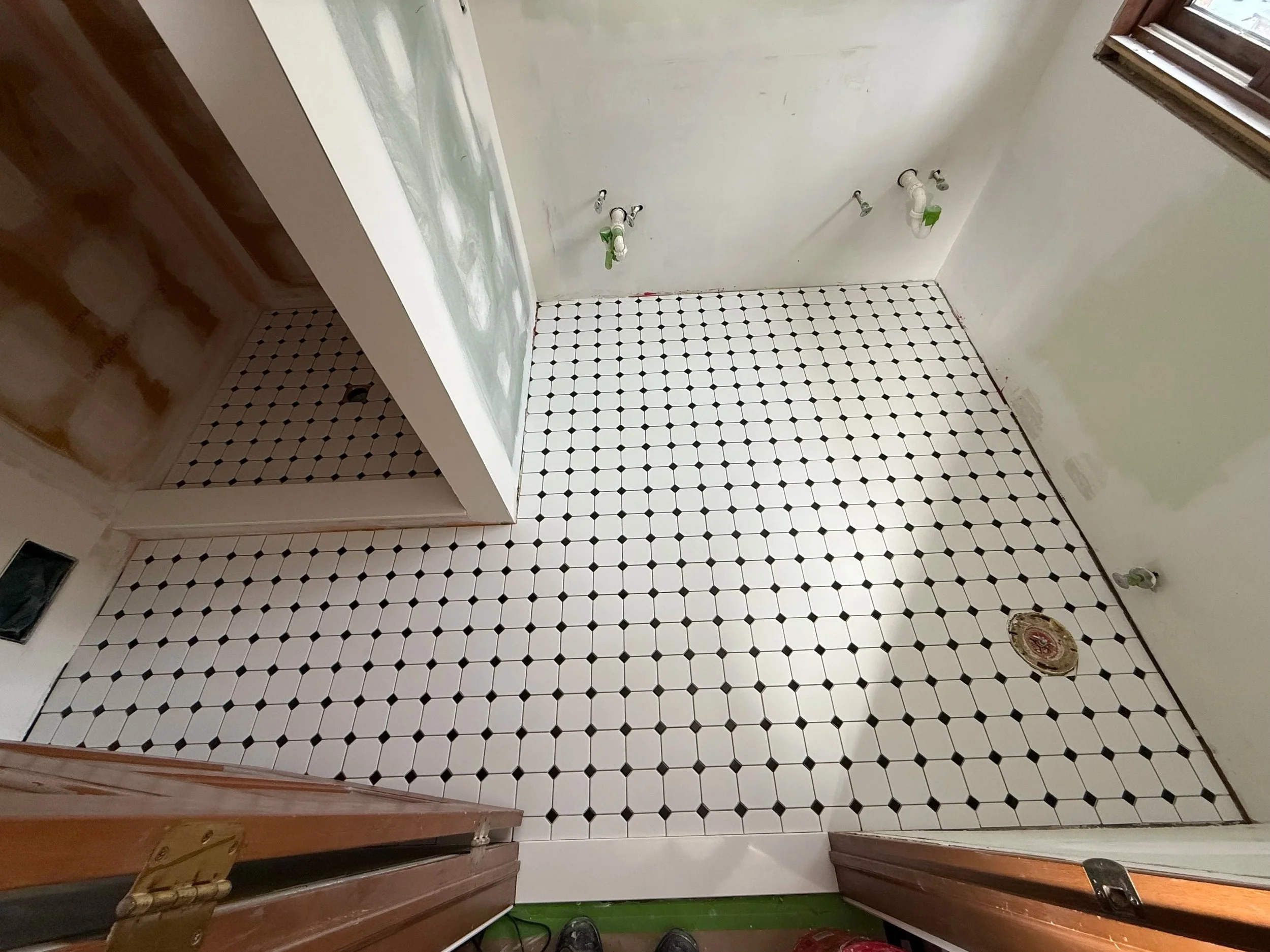 A room under renovation with black and white checkered tile flooring, multiple plumbing pipes and fittings on the wall, a floor drain, and unfinished walls with patches of paint and drywall.