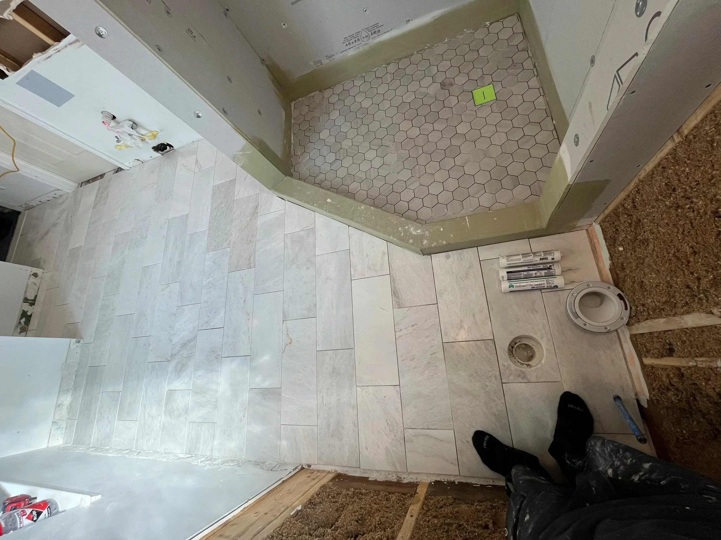 Interior view of a bathroom under renovation with tiled floors and an alcove shower area with hexagonal tiles. Construction supplies like tubes of sealant and a pipe are visible on the floor. The person's feet are visible at the bottom of the image.
