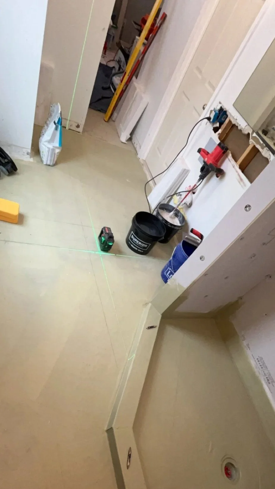 A construction or renovation scene with laser level lines projected on the floor, tools such as a drill, bucket, and level, and a partially finished wall with an open electrical box.