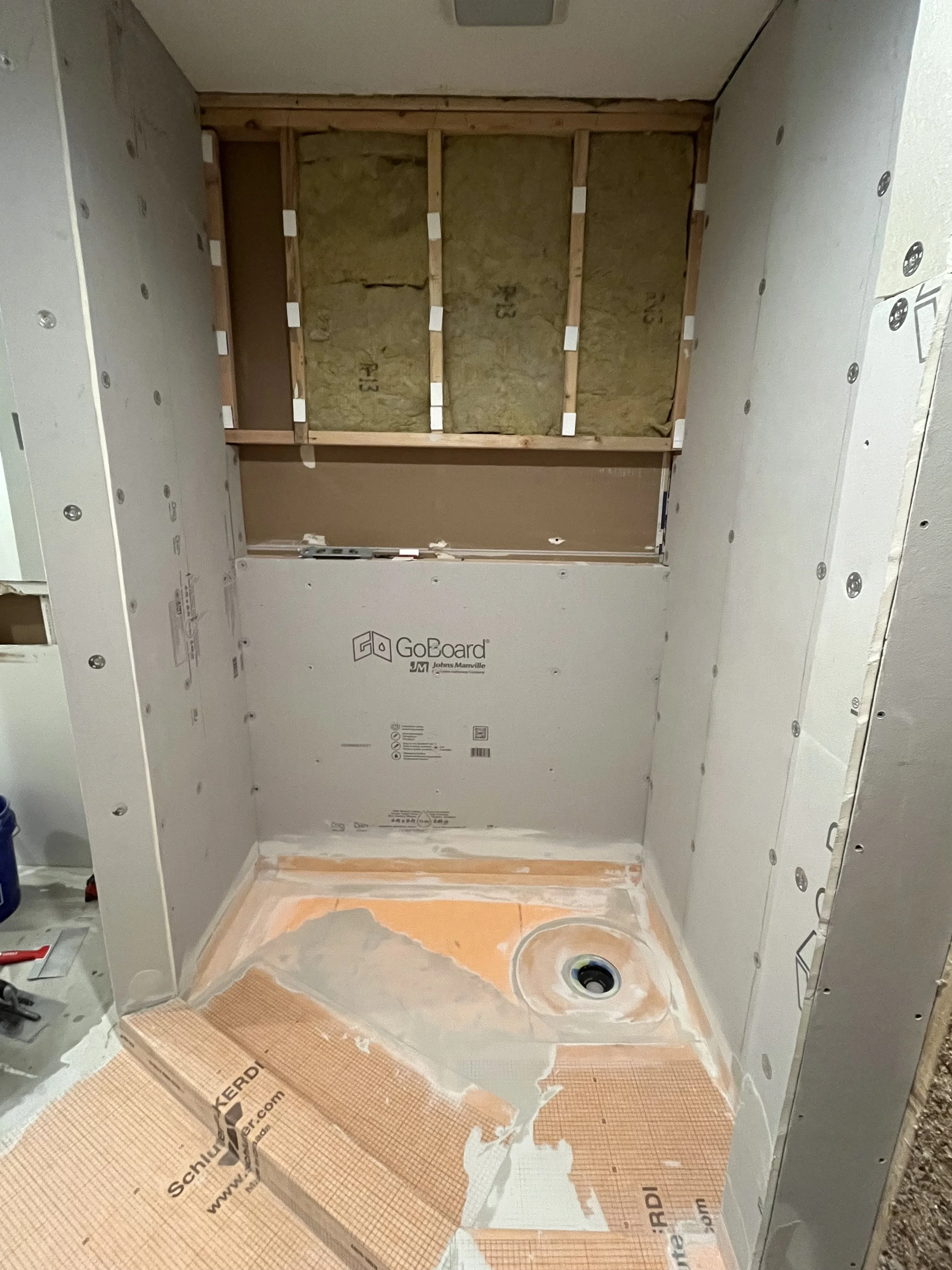 Bathroom shower area under construction with drywall and insulation, with a drain installed in the floor and a partially tiled floor.