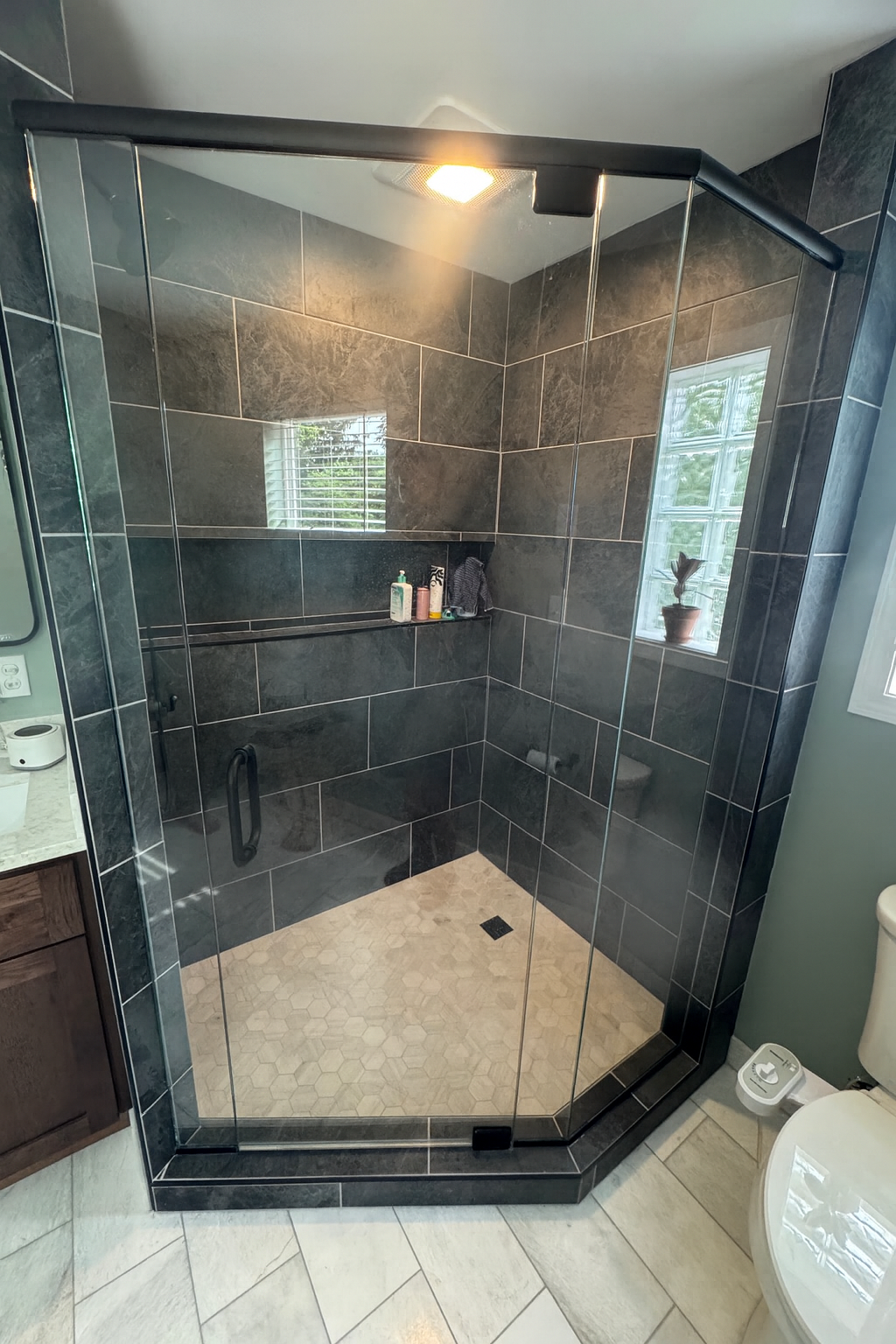 A modern bathroom with a walk-in glass shower featuring dark gray tiled walls and a hexagonal beige tiled floor. The shower has a built-in shelf with toiletries, a window with blinds, and a black shower rod. Adjacent to the shower, part of a wooden v