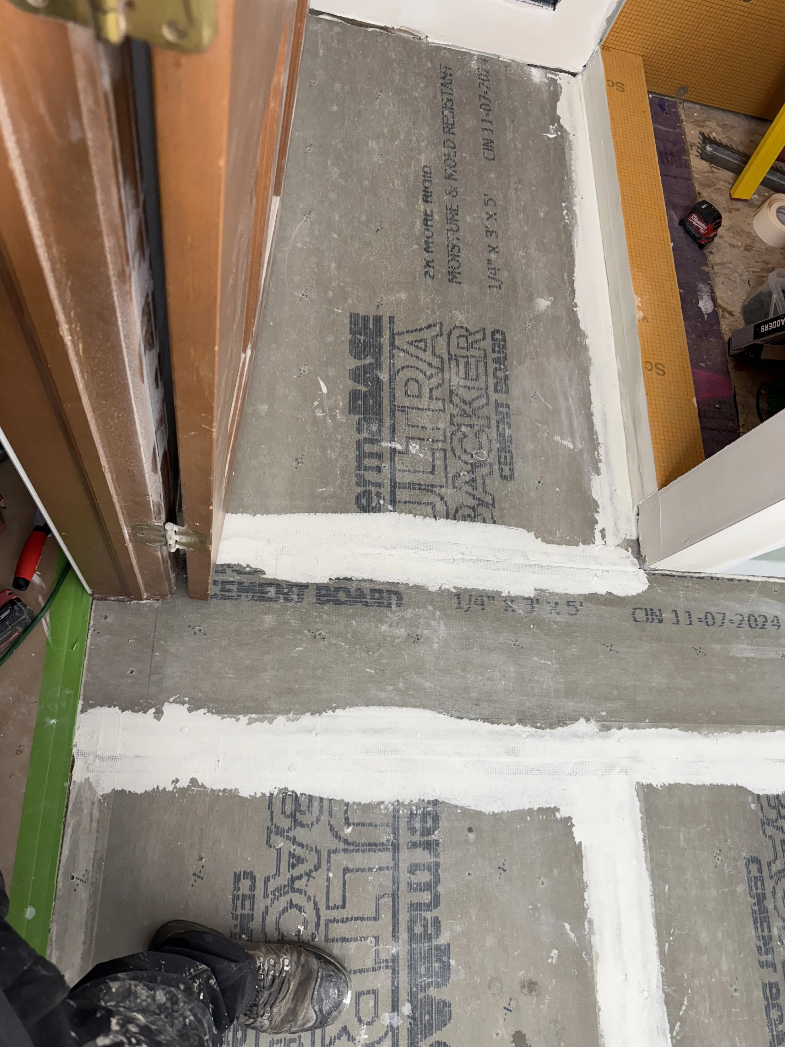 Floor under renovation showing cement subfloor with markings and white patching compound, door frame on the left, and construction tools and supplies on the right.