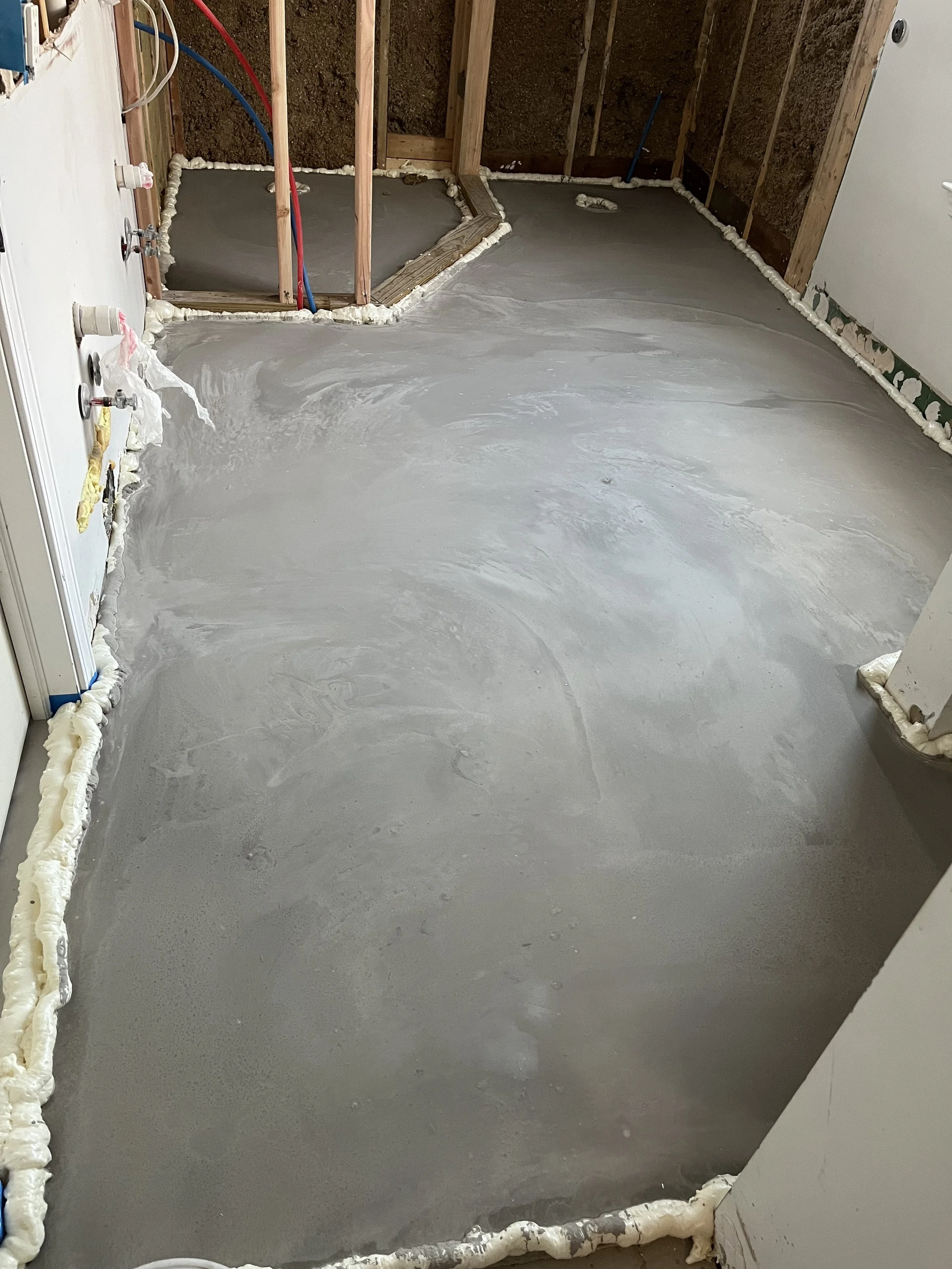 Newly poured concrete floor in a bathroom under construction, with plumbing and framing visible.