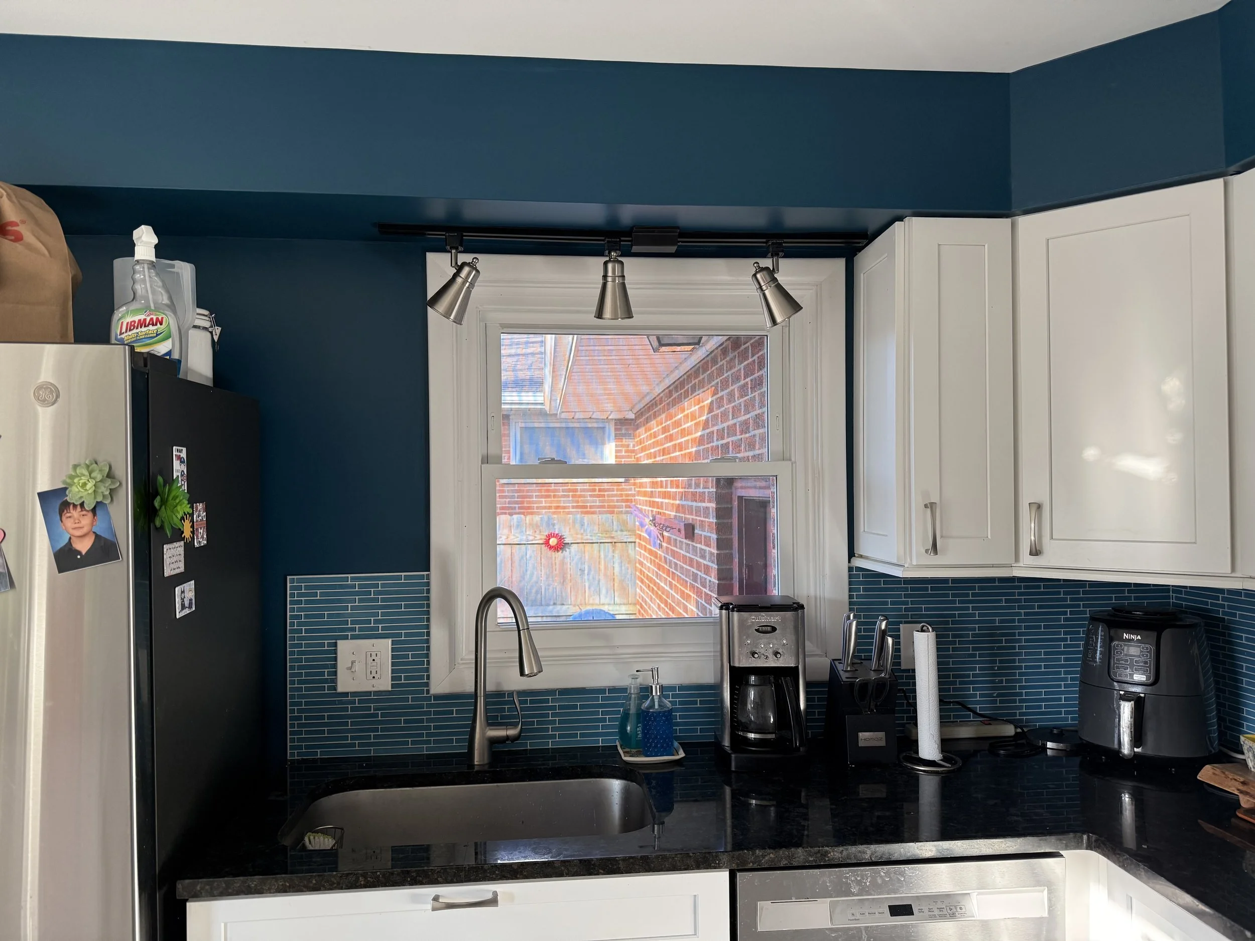 Kitchen with a window above the sink, white cabinets, a blue tile backsplash, a black countertop, and various appliances including a coffee maker and air fryer.
