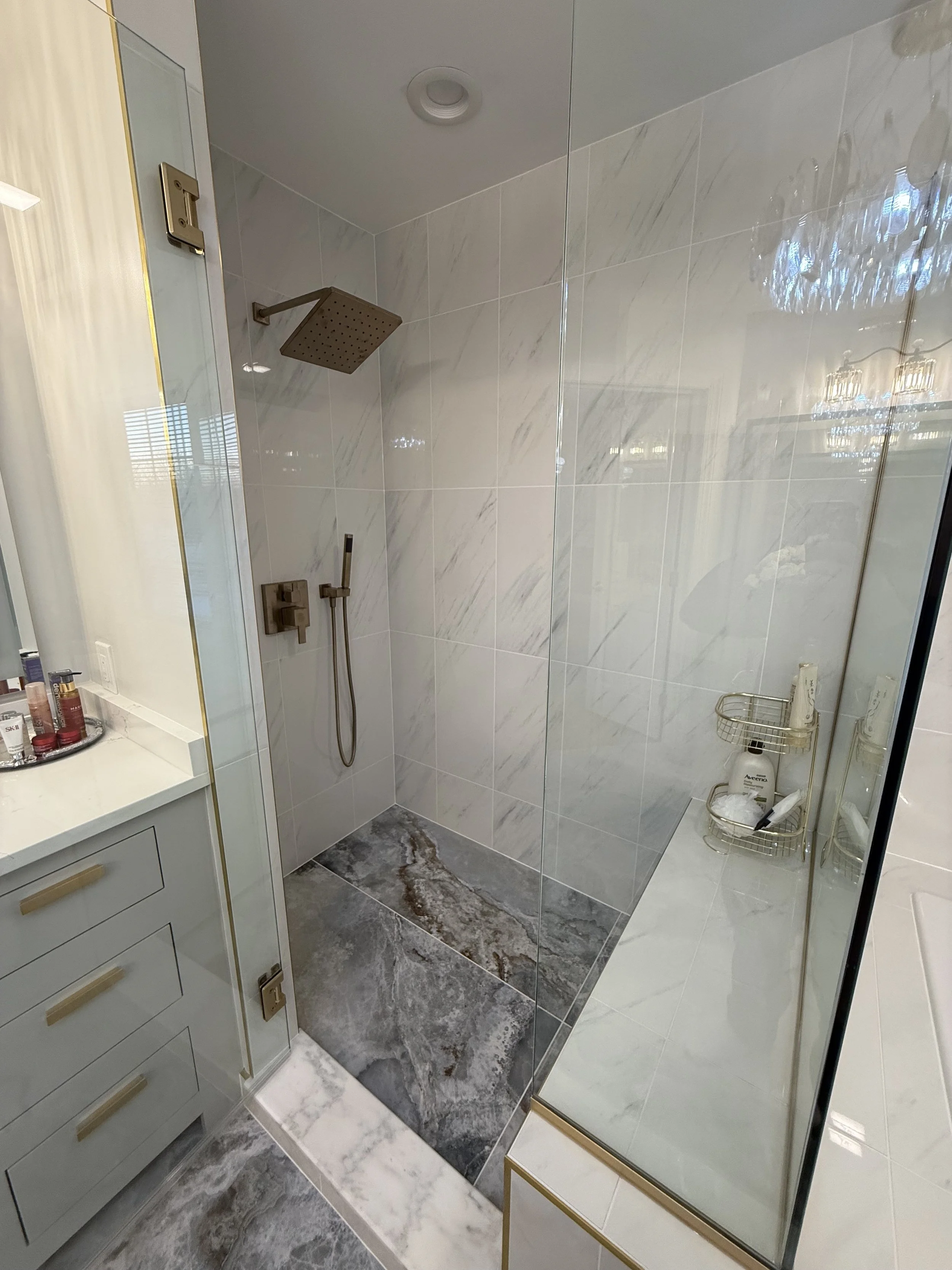 Modern bathroom with walk-in shower featuring large white marble tiles and a large rain shower head. There is a glass shower enclosure, a white vanity with toiletries, and marble flooring.