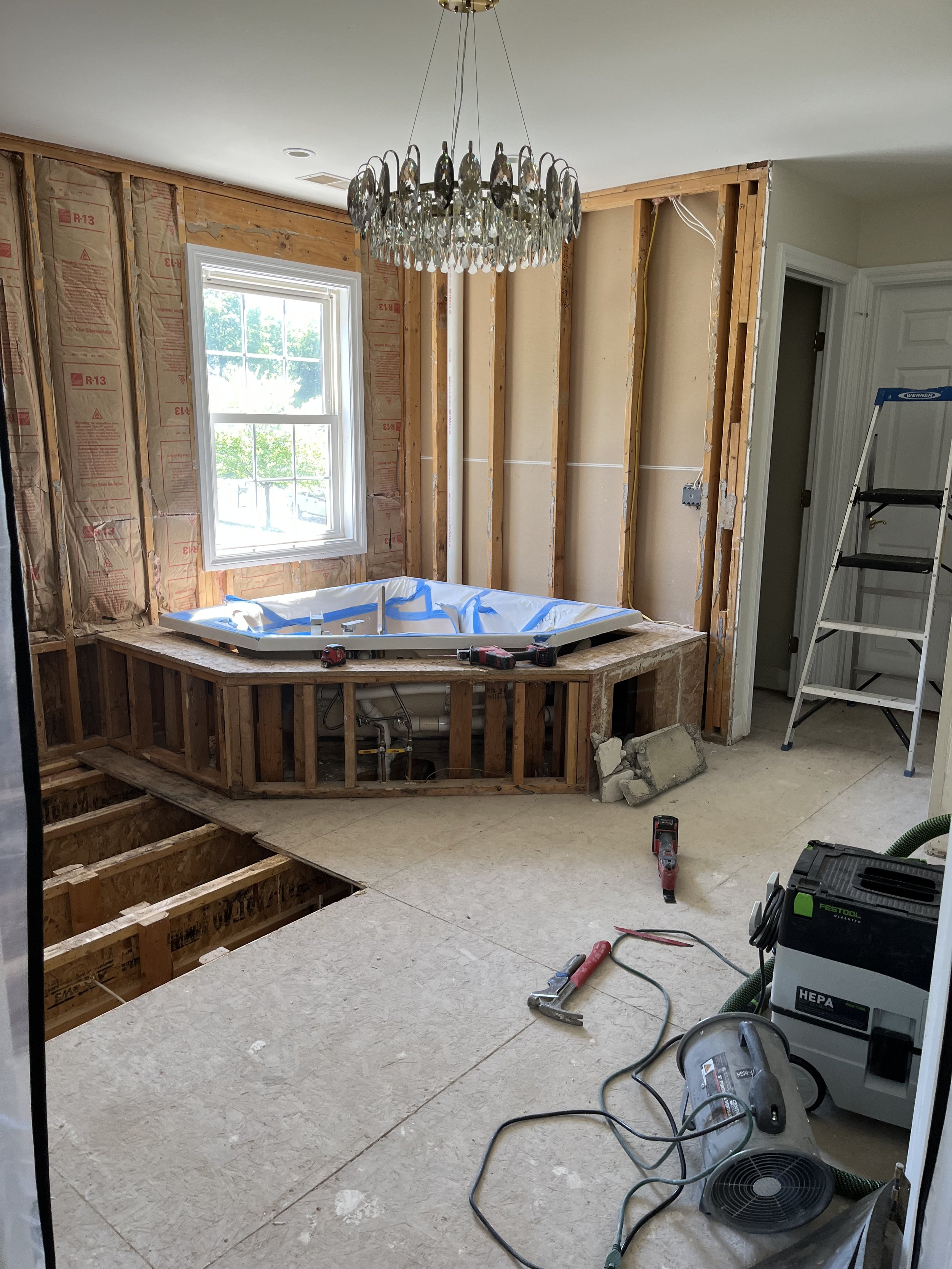 Bathroom renovation in progress with exposed wall framing and a hot tub installed in the corner. Construction tools and materials are scattered around.