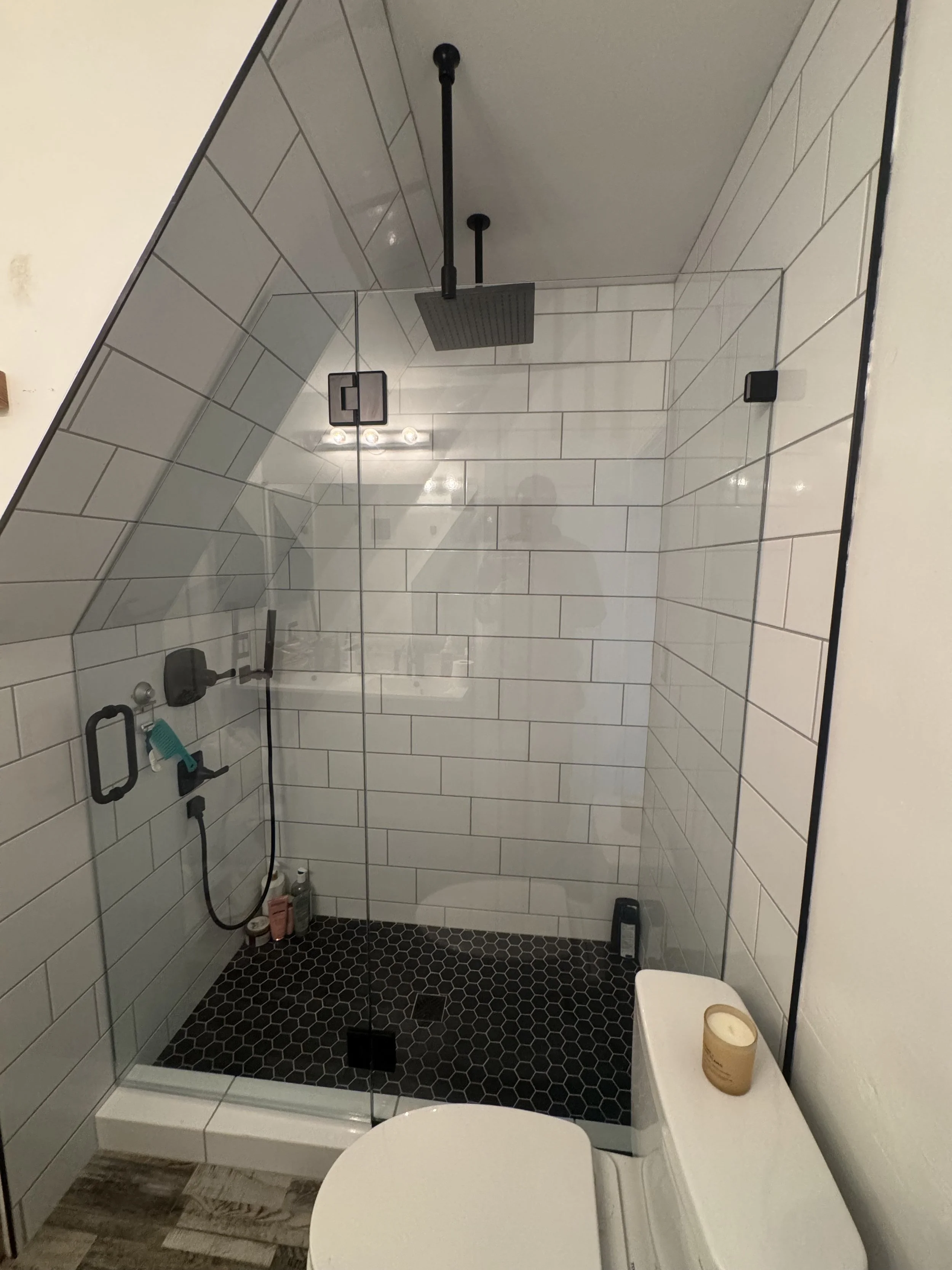 Modern bathroom with a walk-in shower featuring white tile walls, a black hexagonal tile floor, and a ceiling-mounted black rainfall showerhead. A glass partition separates the shower from the toilet, which has a candle on top.