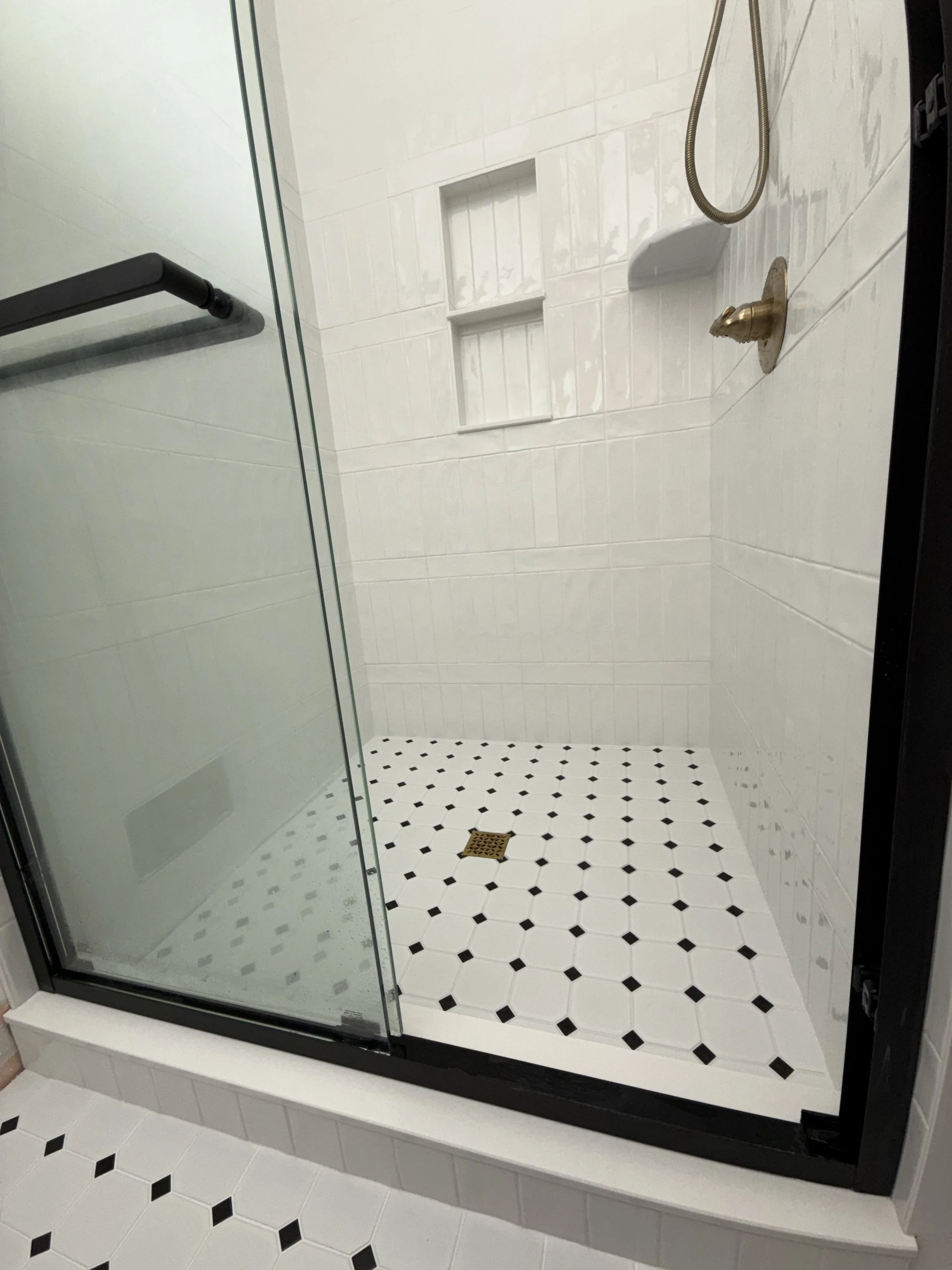 Empty shower stall with black and white patterned floor tiles, built-in shelves on the wall, and a metal shower hook hanging from the wall.
