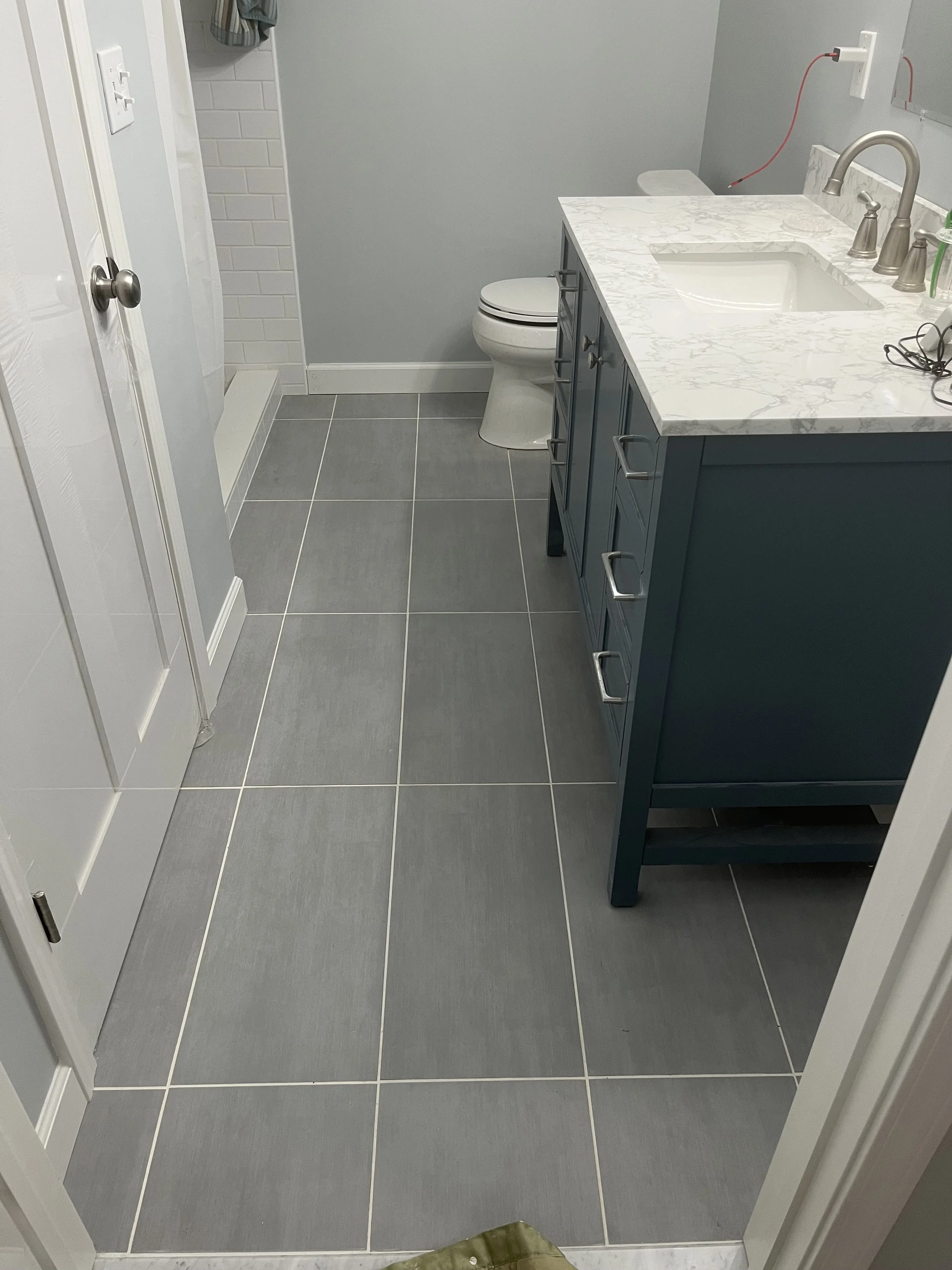 A modern bathroom with gray floor tiles, a blue vanity with a white marble countertop, a white undermount sink, a silver faucet, a toilet, and a shower area with white subway tile walls. There are toiletries and a towel on a hook.