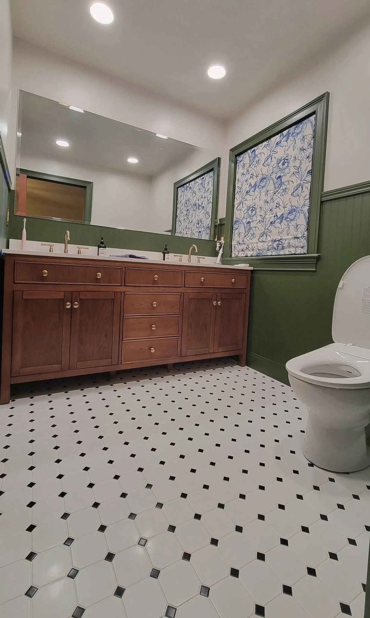 Bathroom with a wooden vanity, a large mirror, green walls, patterned window shades, a toilet, and a black-and-white hexagonal tile floor.